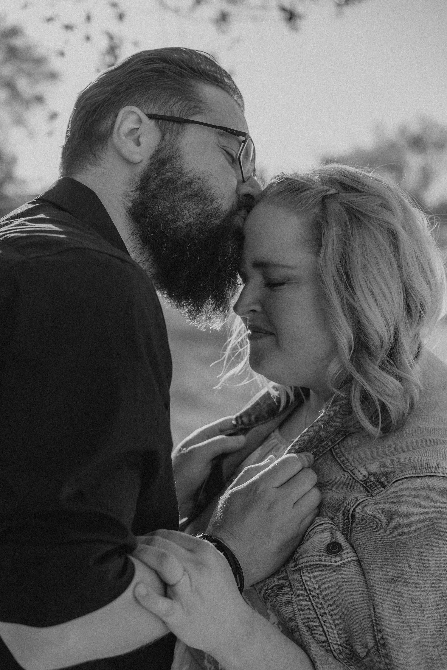 Black and white photo of a bearded man kissing a smiling blonde woman outdoors in a romantic couple portrait.