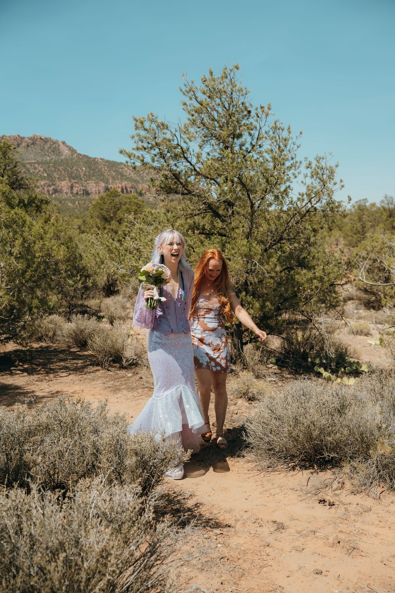 Two women walking on a desert trail, one in a blue dress holding flowers, surrounded by shrubs and red rock mountains.
