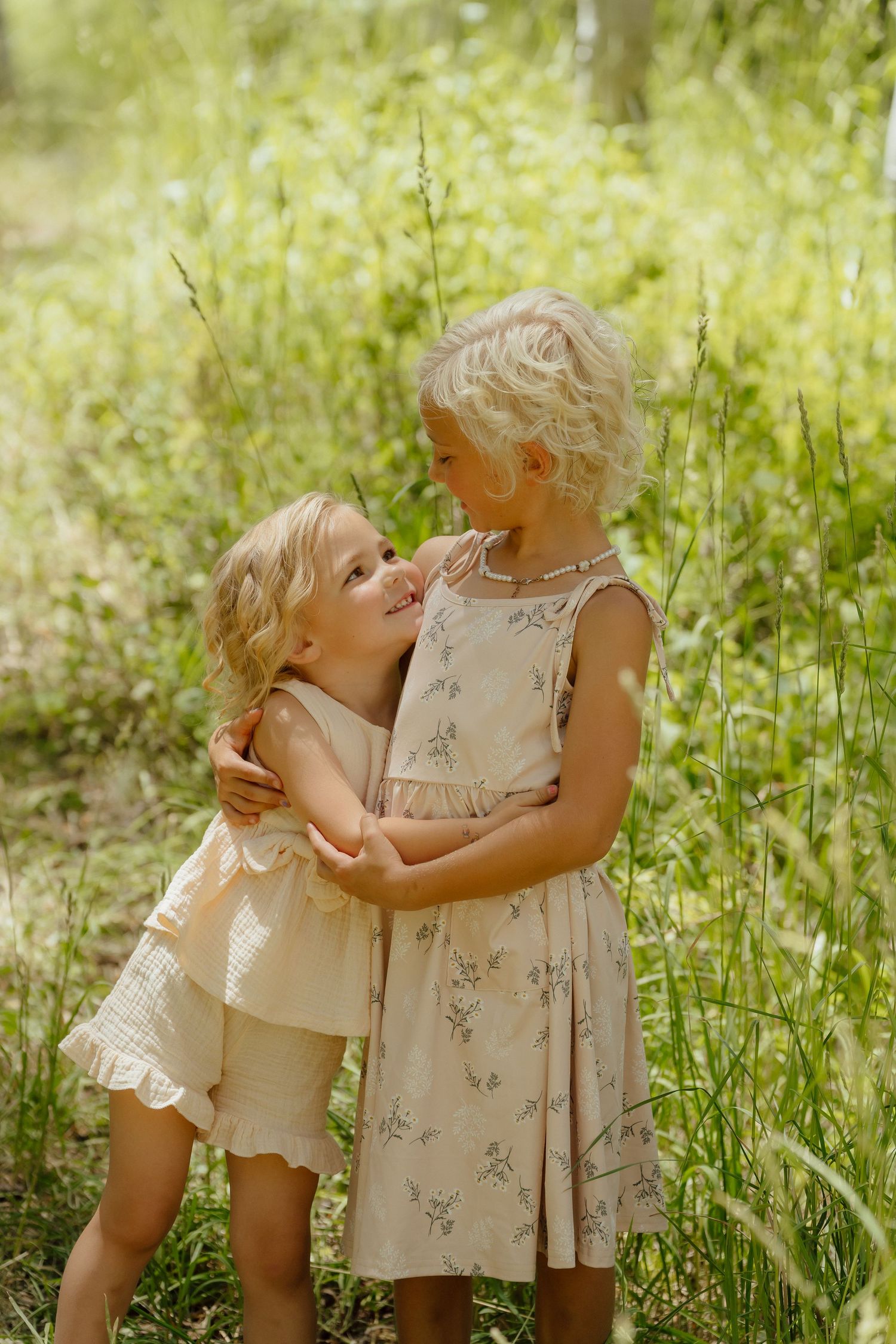 Two young blonde girls in floral dresses embracing outdoors in a sunny green meadow.