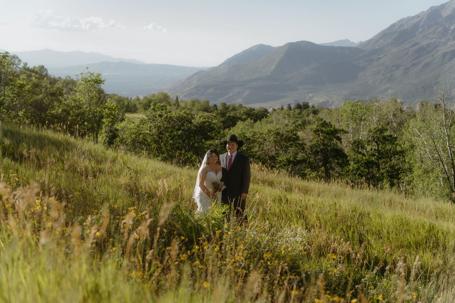 A bride and groom embrace on a lush green hillside with stunning mountain views in the background.