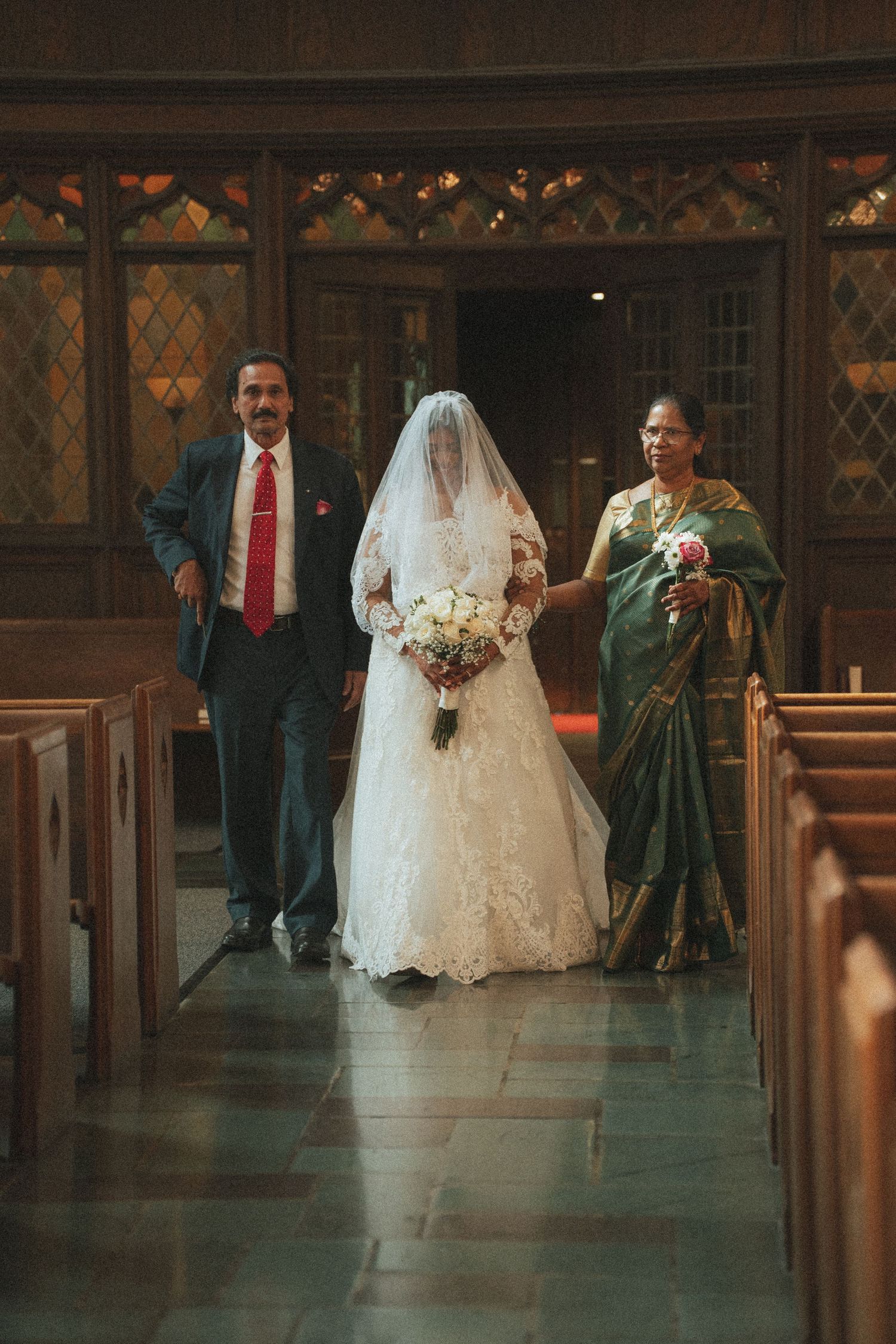 Bride walks down church aisle with parents, wearing lace gown and veil, holding bouquet, in wood-paneled chapel.