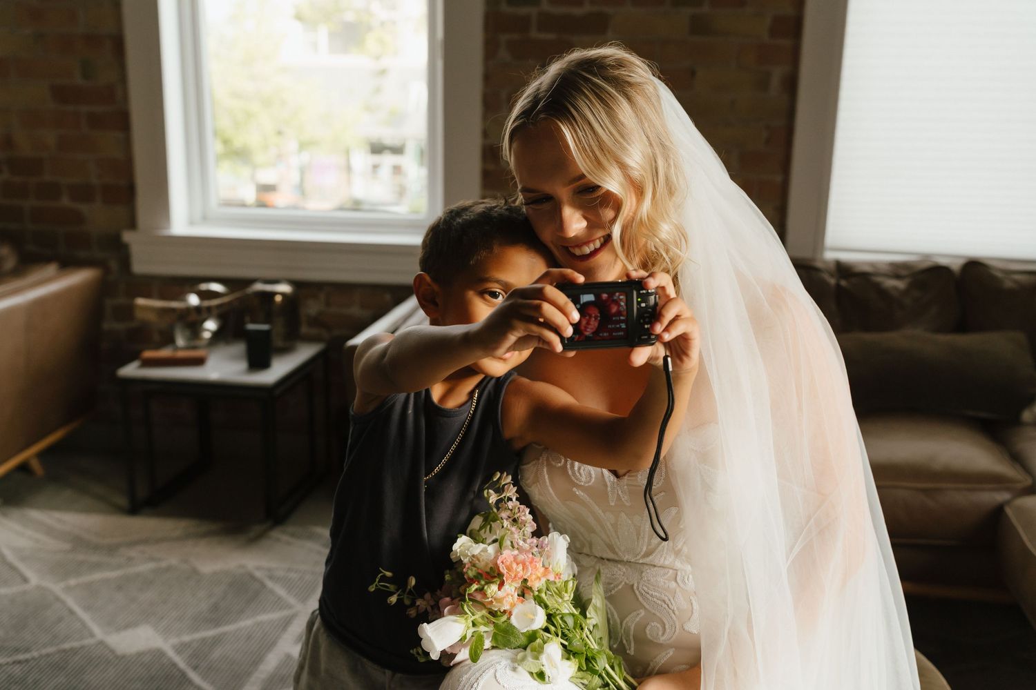 Bride in white gown and veil takes a photo with a child holding a floral bouquet indoors near a window.