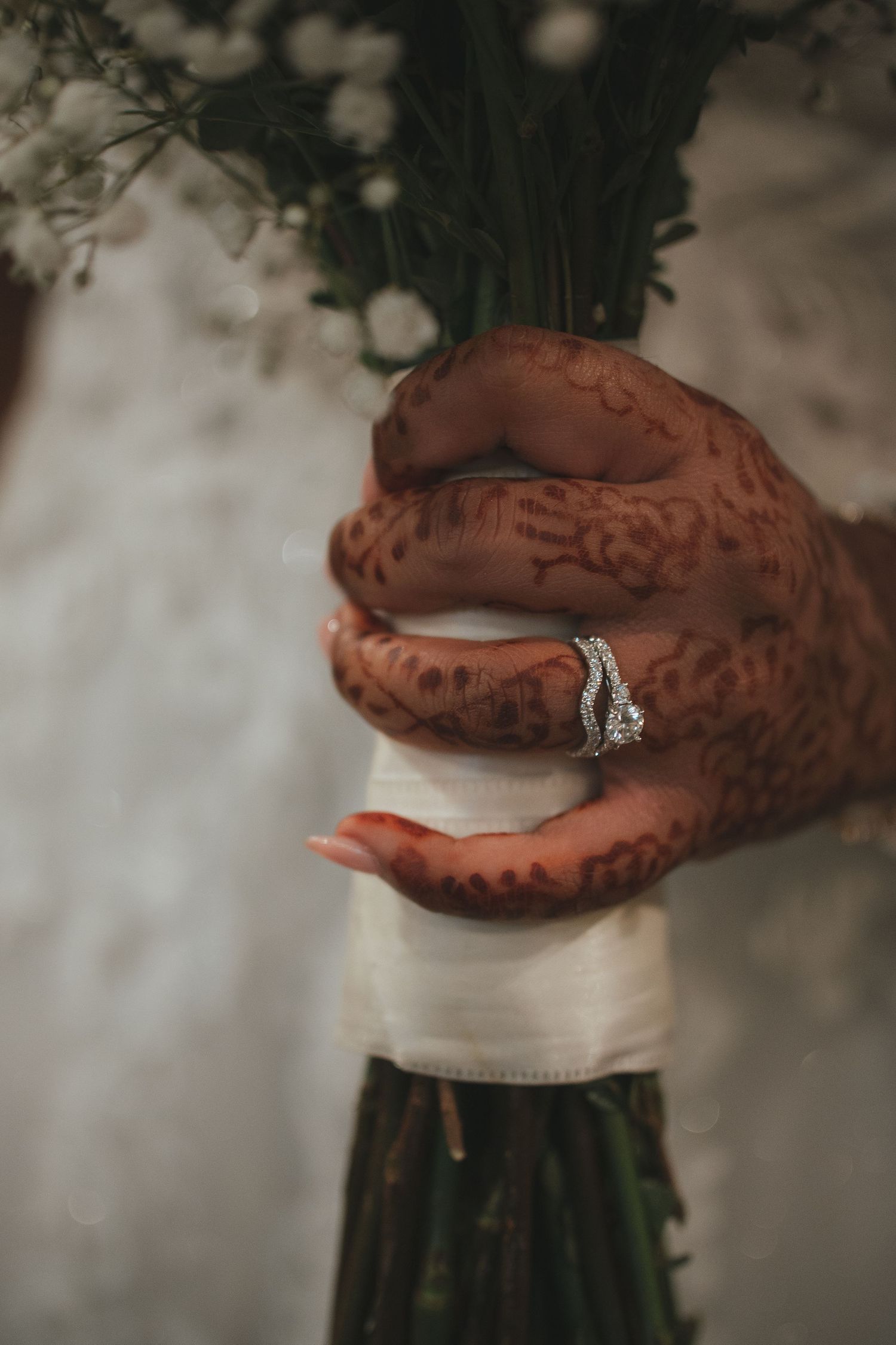 Close-up of a bride's henna-adorned hand holding a white floral bouquet, wearing a diamond ring.