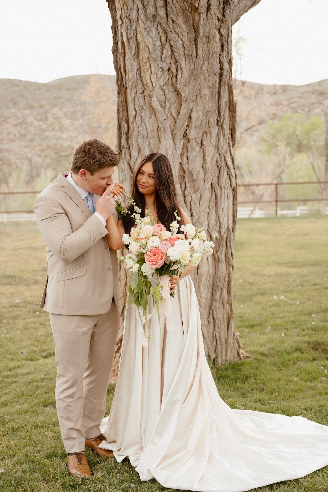 Groom in tan suit kisses brides hand as she holds pink and white bouquet by large tree outdoors.