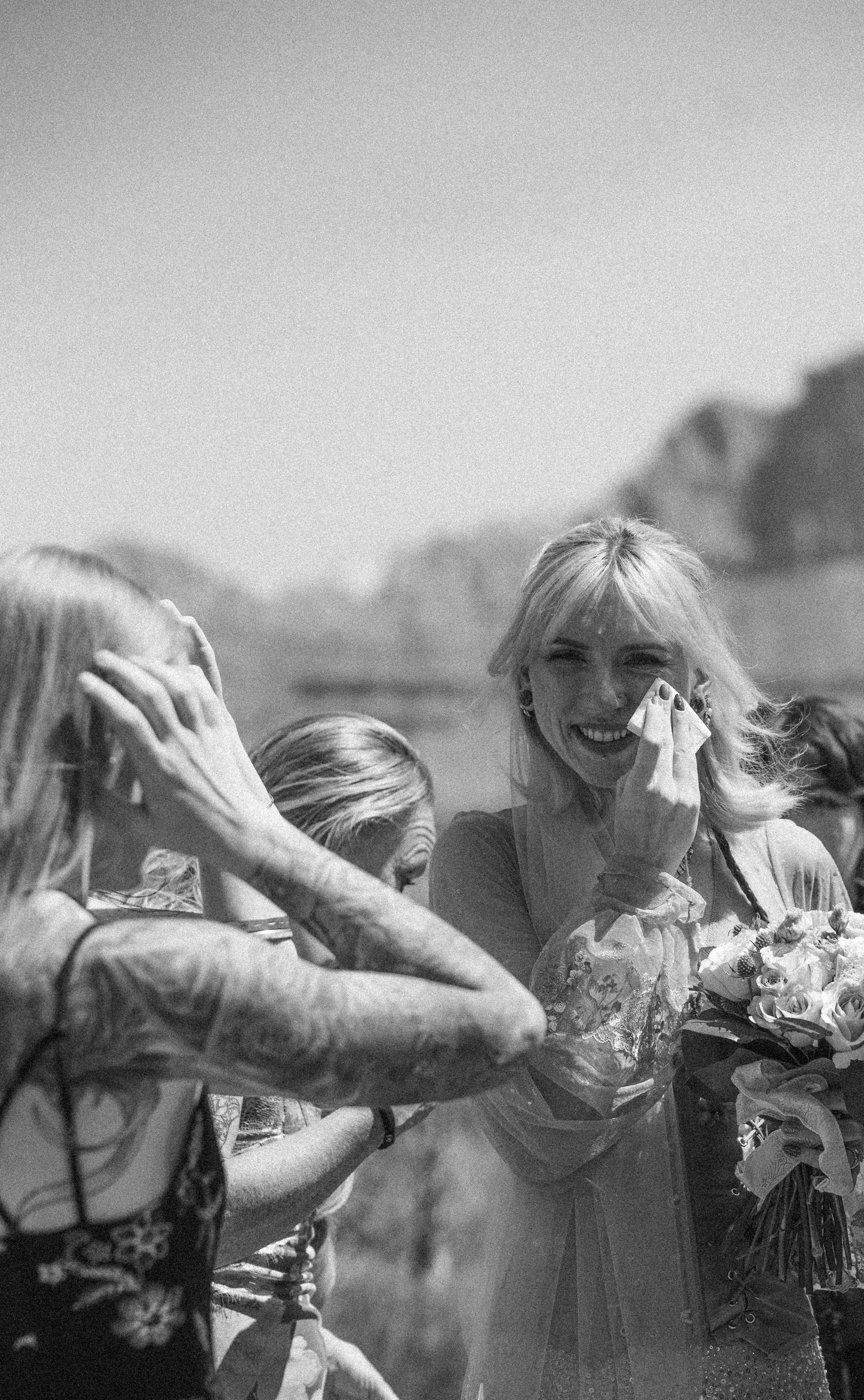 Black and white photo of emotional bride wiping tears with tissue, holding bouquet, surrounded by guests at outdoor wedding.