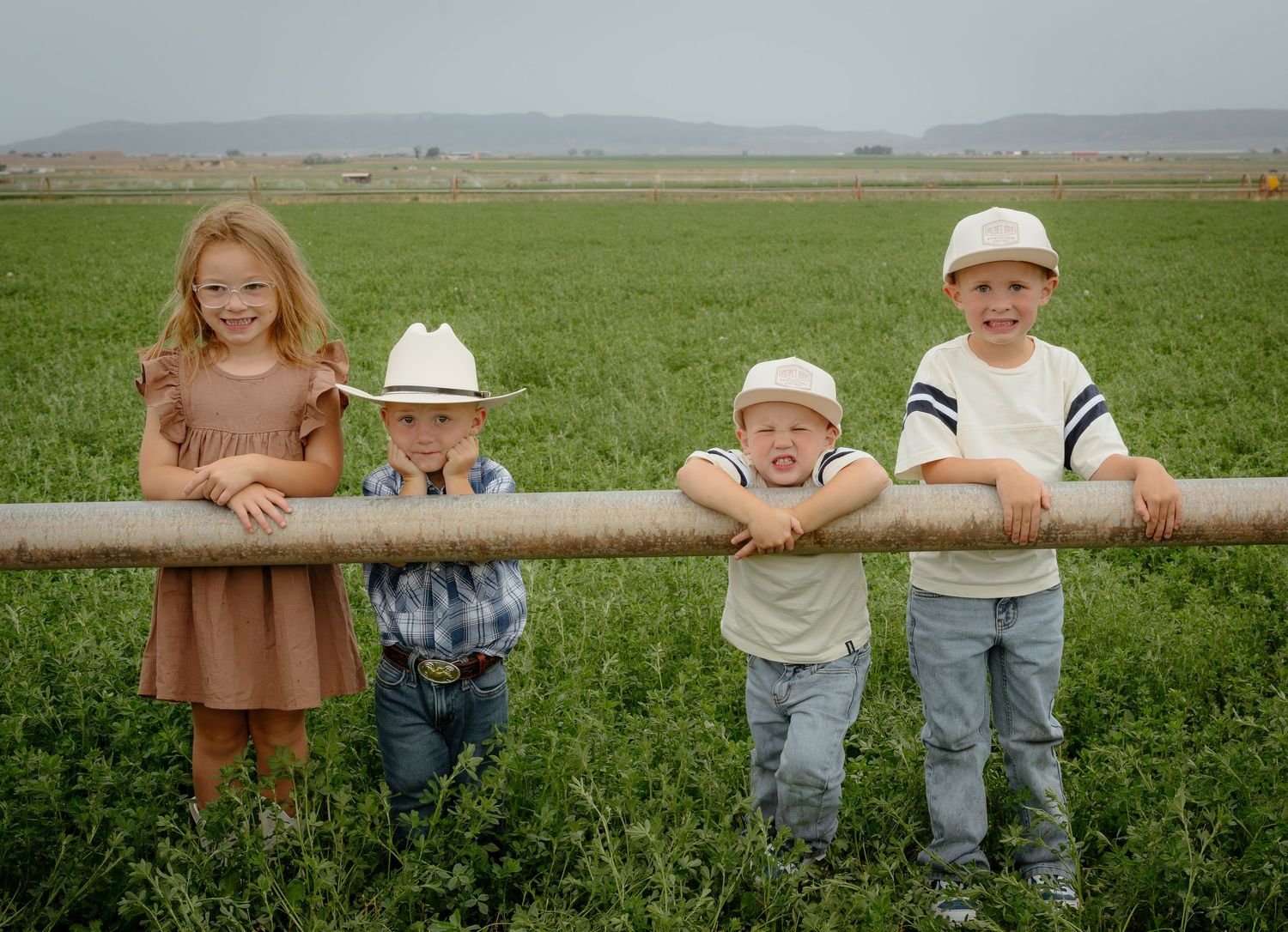 Four young children lean on a rustic fence rail in a green farm field, two wearing cowboy hats, smiling outdoors.