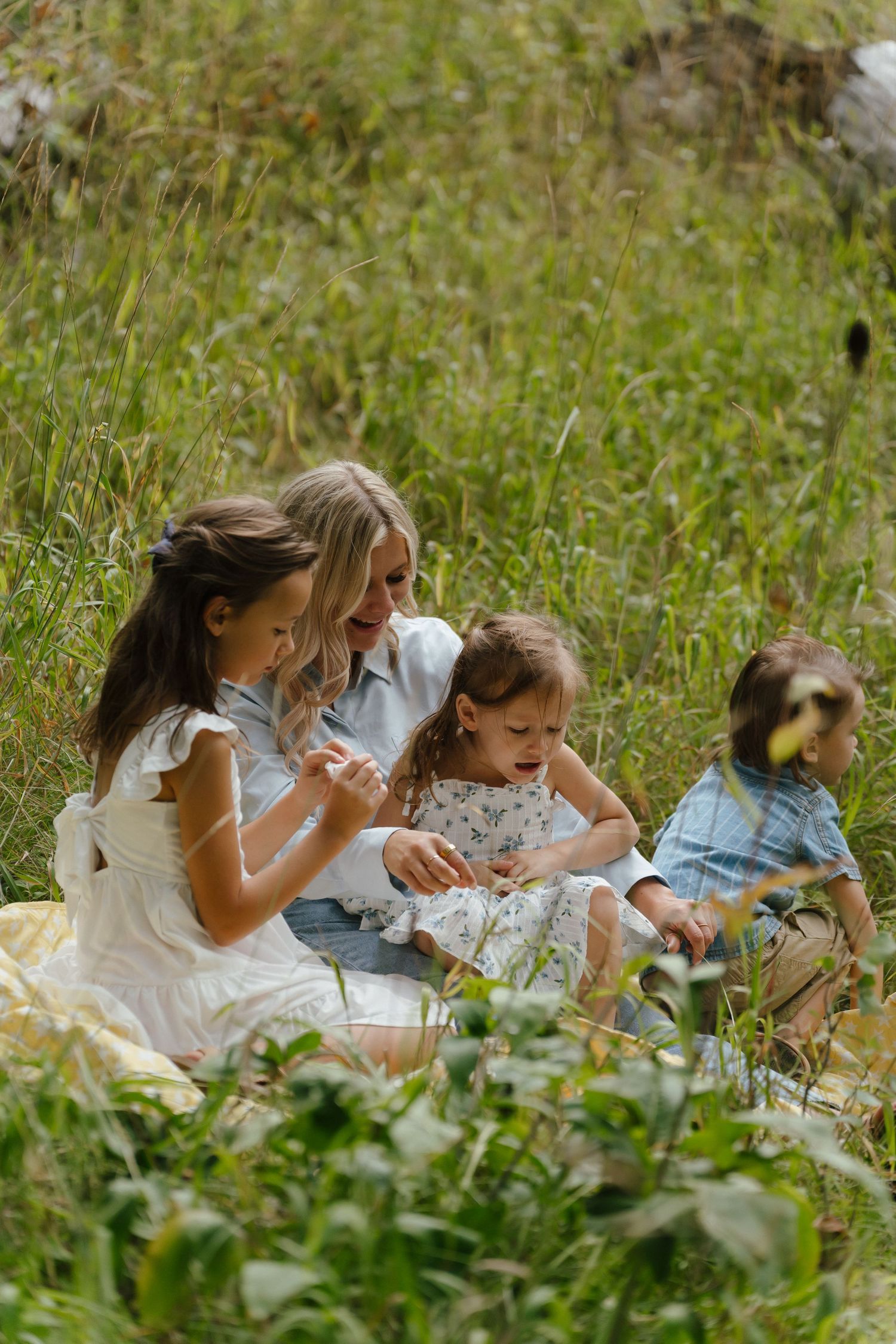 A mother sits in a green meadow with her three young children, sharing a joyful moment together outdoors.