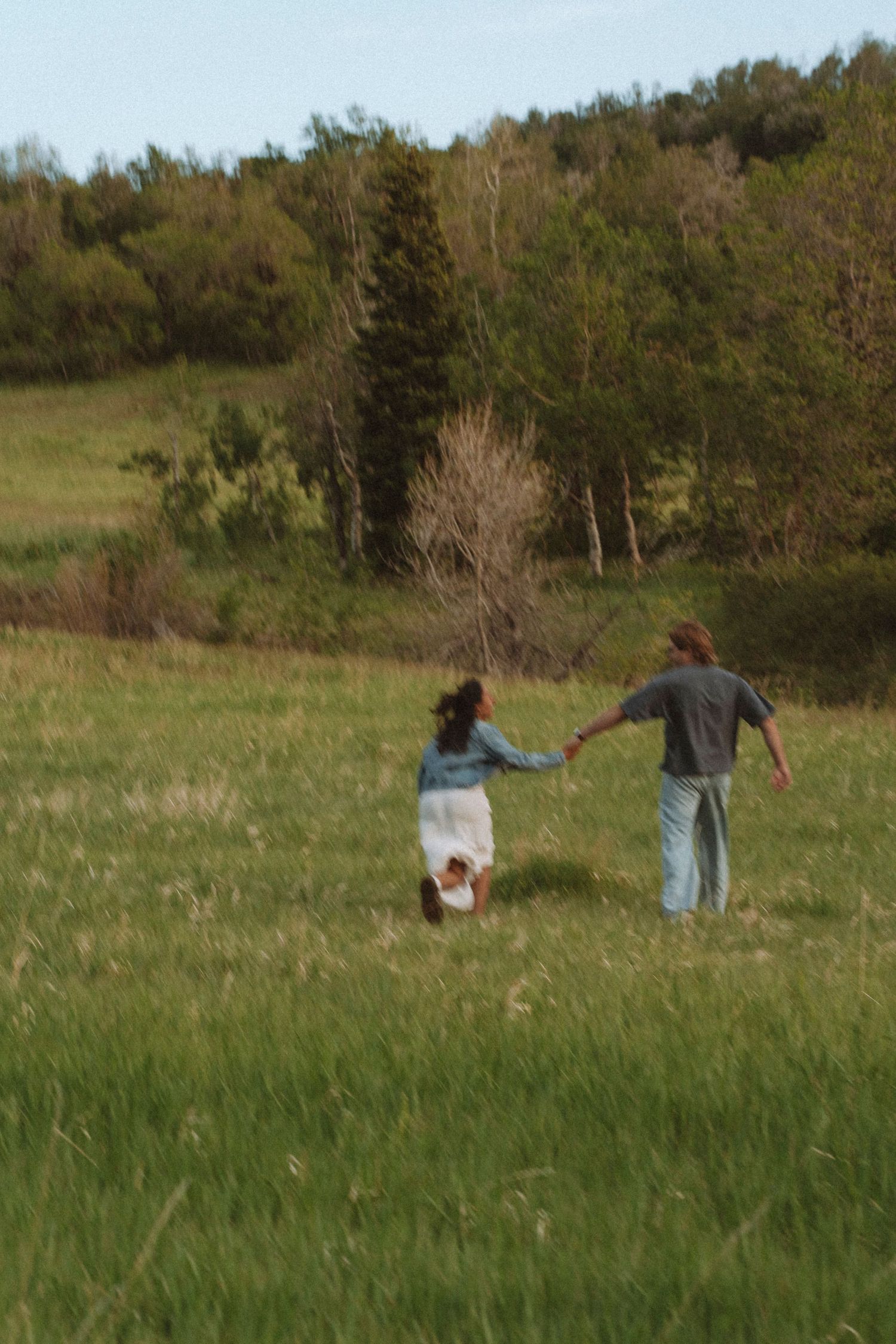 A couple holds hands while running through a lush green meadow with trees and hills in the background.