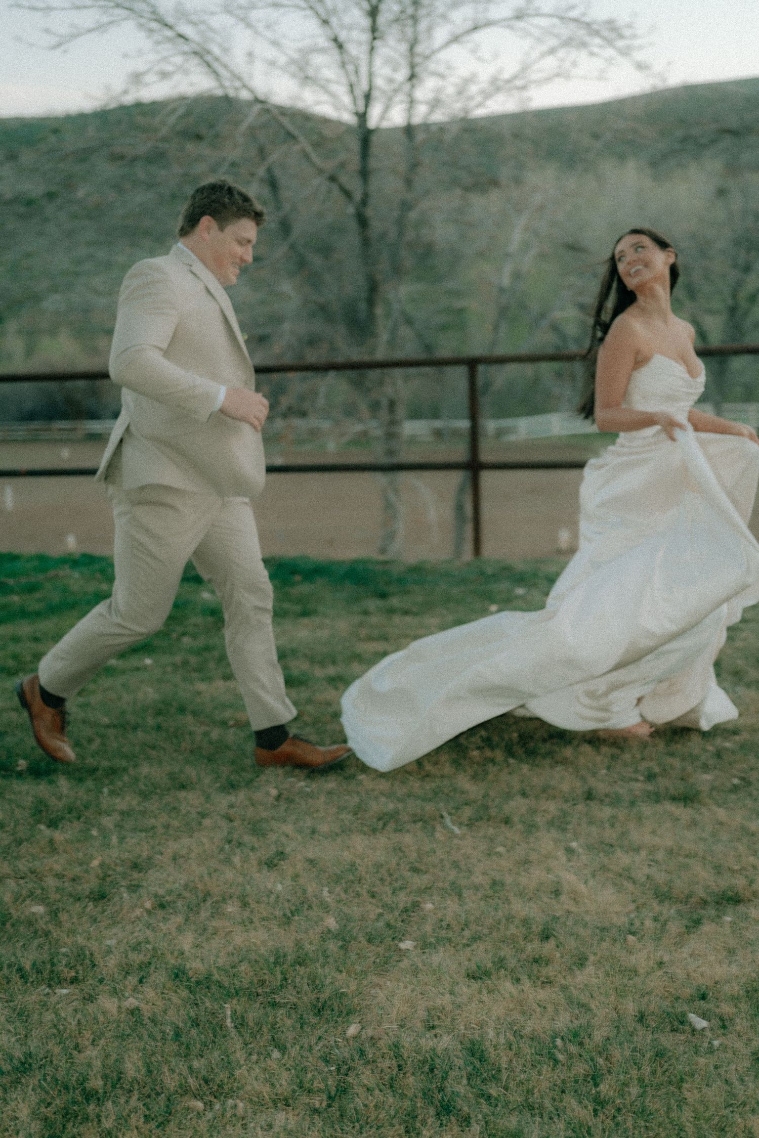 A joyful bride in a flowing white gown and groom in a tan suit run together outdoors on a grassy field.