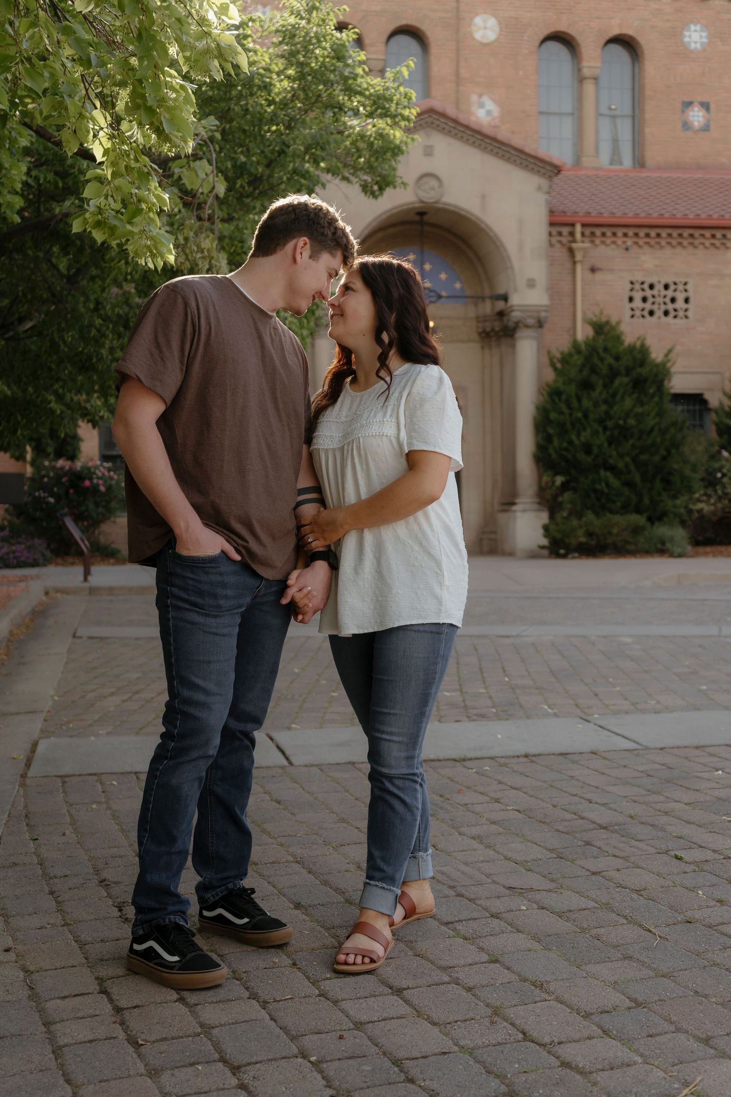 A young couple shares a romantic moment holding hands outside a brick building with arched windows and lush green trees.