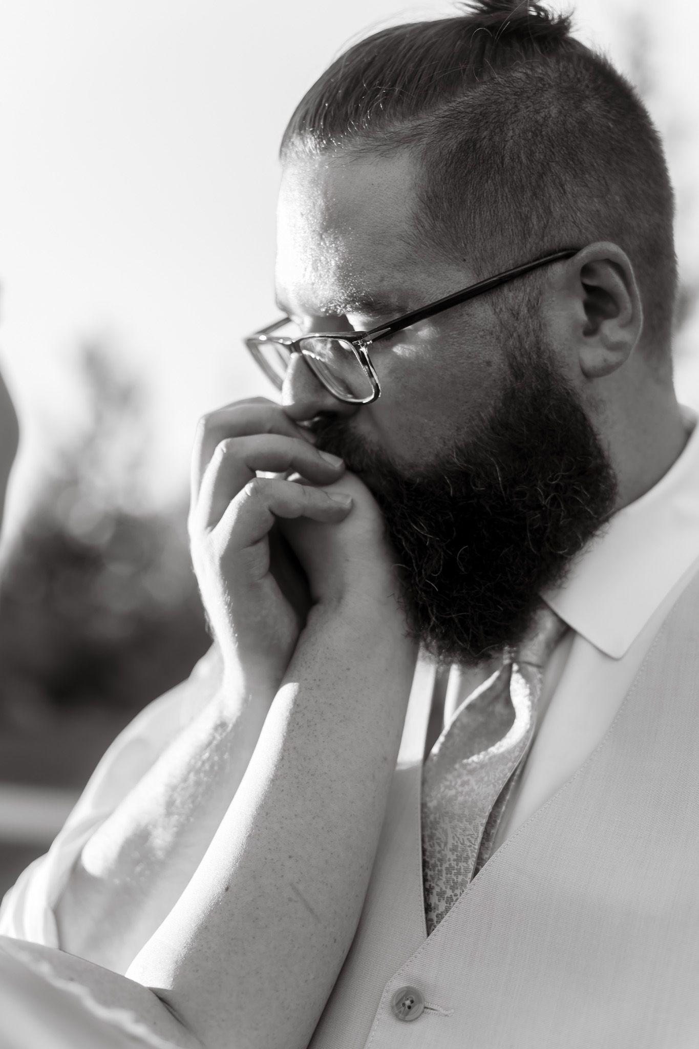 Black and white portrait of a bearded man with glasses and a bun kissing a hand, wearing a white shirt.