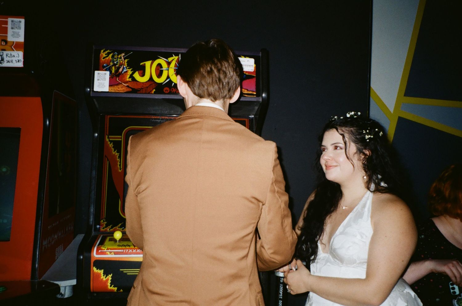 Bride wearing wedding dress admiring groom in tan suit playing arcade pinball machine game at Player 2 Arcade Bar in Madison, Wisconsin