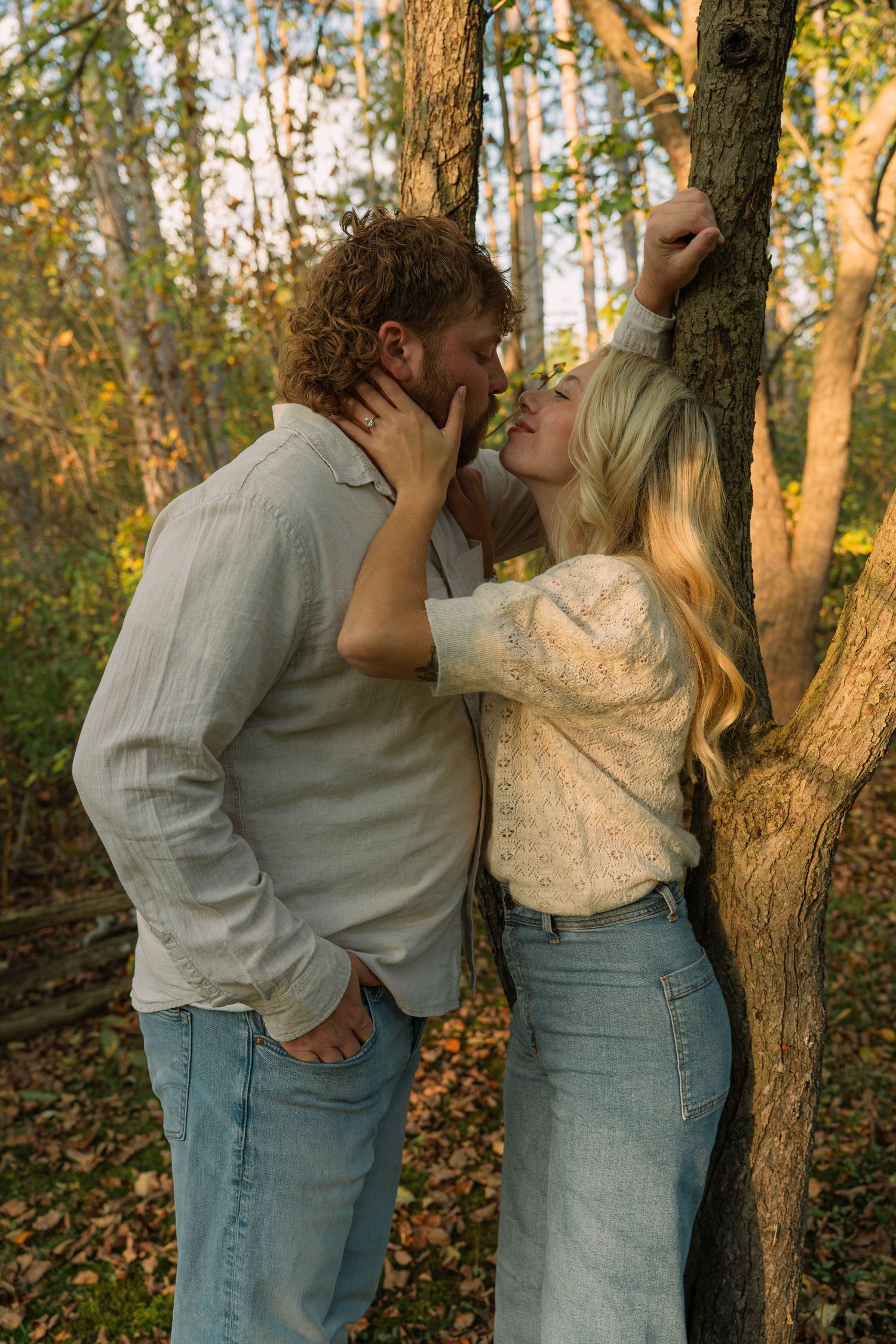 man and woman kissing against tree in woods wearing blue jeans and white shirt with blonde hair