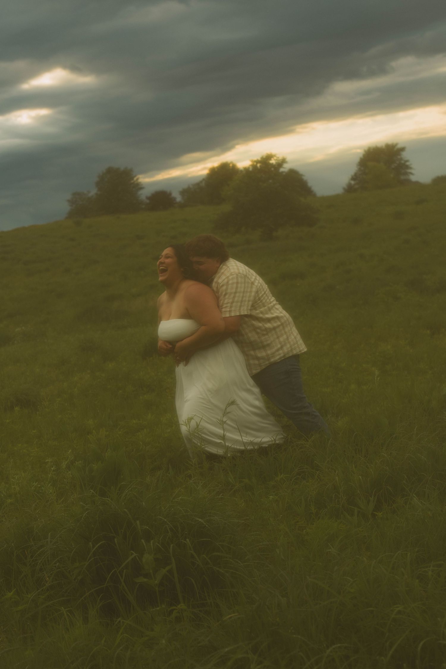 laughing couple in tall grassy field and overcast sky in big embrace with hazy filter over the image