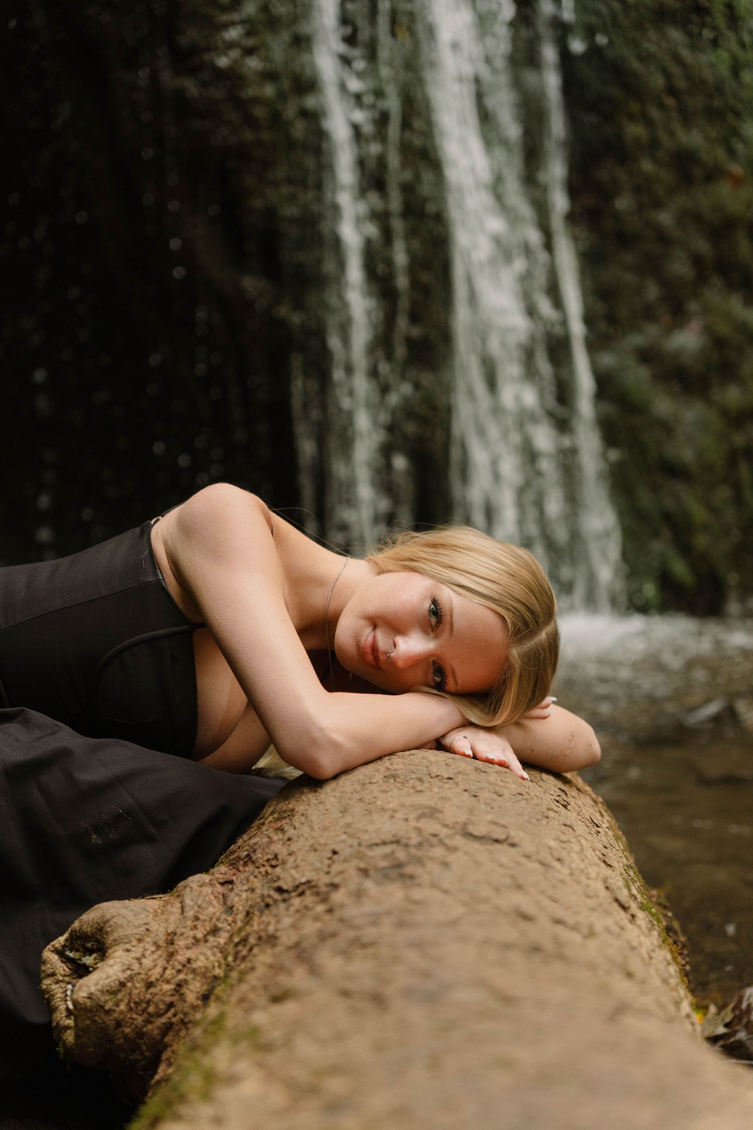 blonde girl laying on tree branch with waterfall in background wearing black dress for senior photos in Madison, Wisconsin