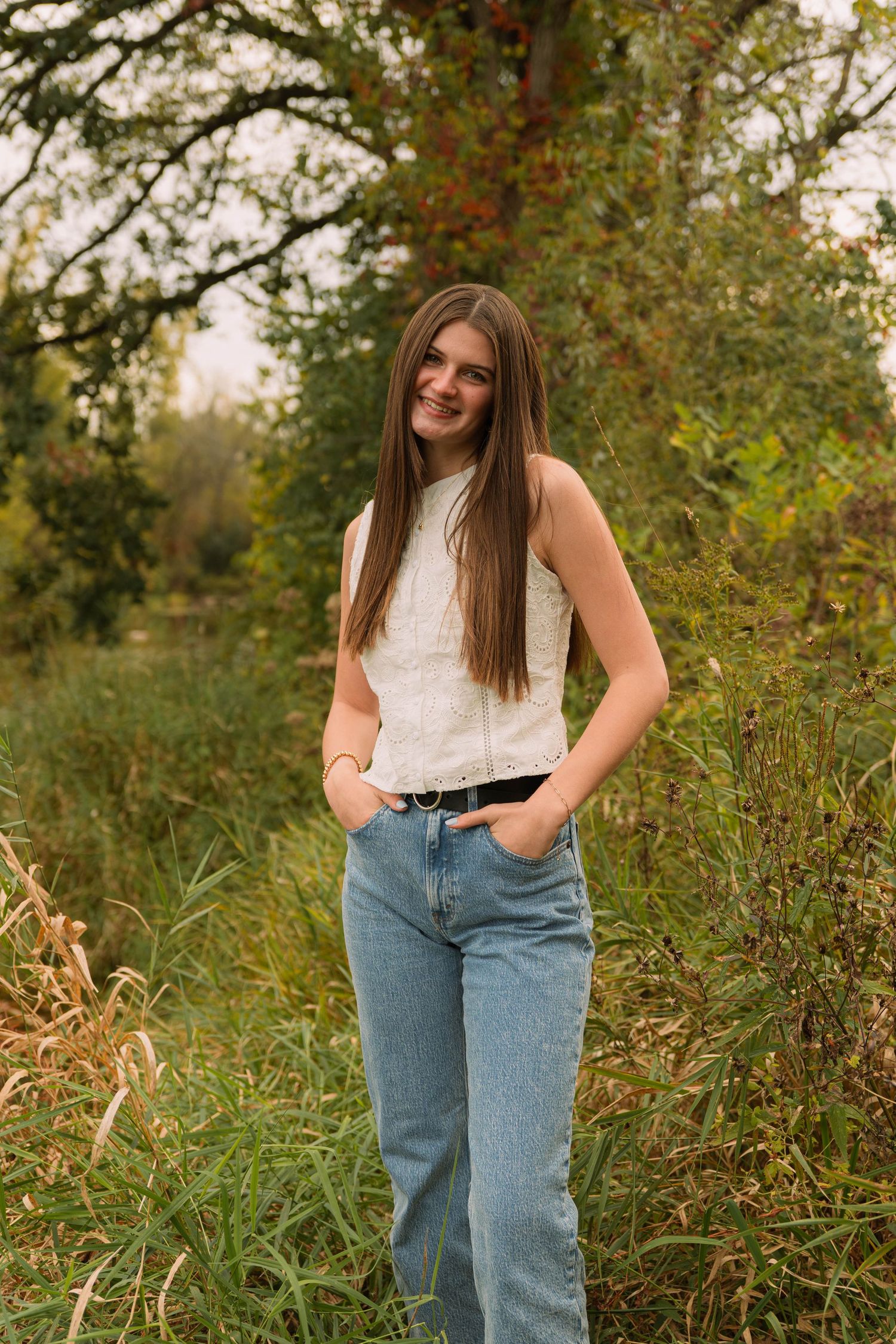 smiling young girl with hands in blue jean pockets in tall green grass and trees with brown hair and white top at Pamperin Park in Green Bay