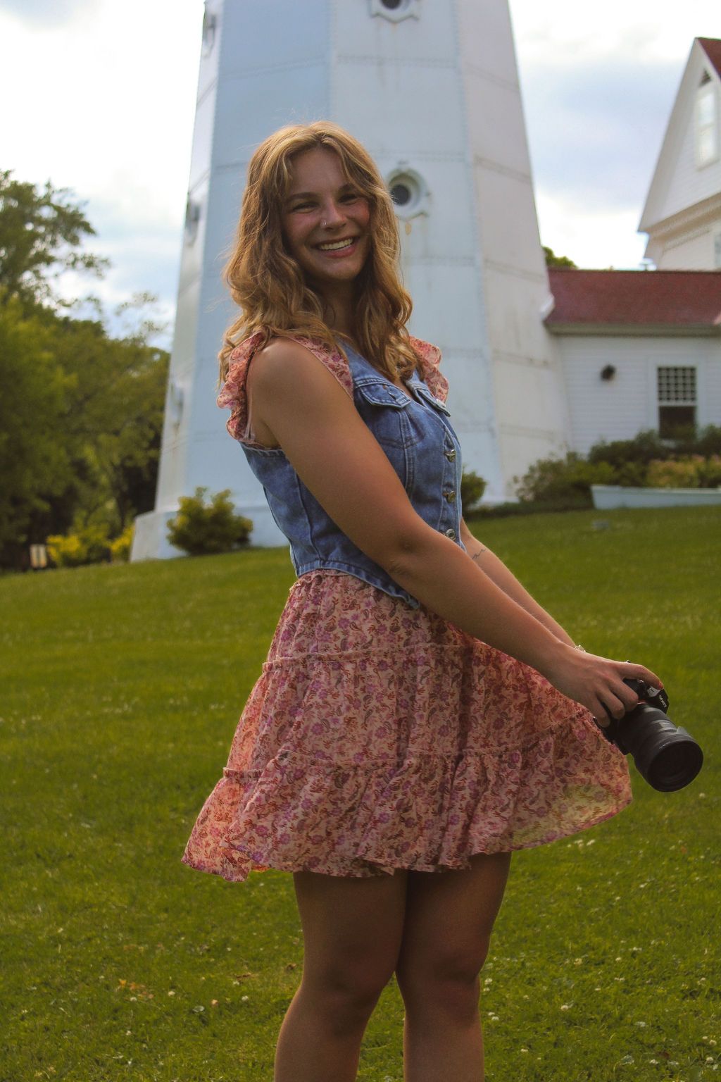 Girl photographer smiling holding camera in front of white lighthouse wearing blue denim vest and pink paisley dress in Milwaukee, Wisconsin
