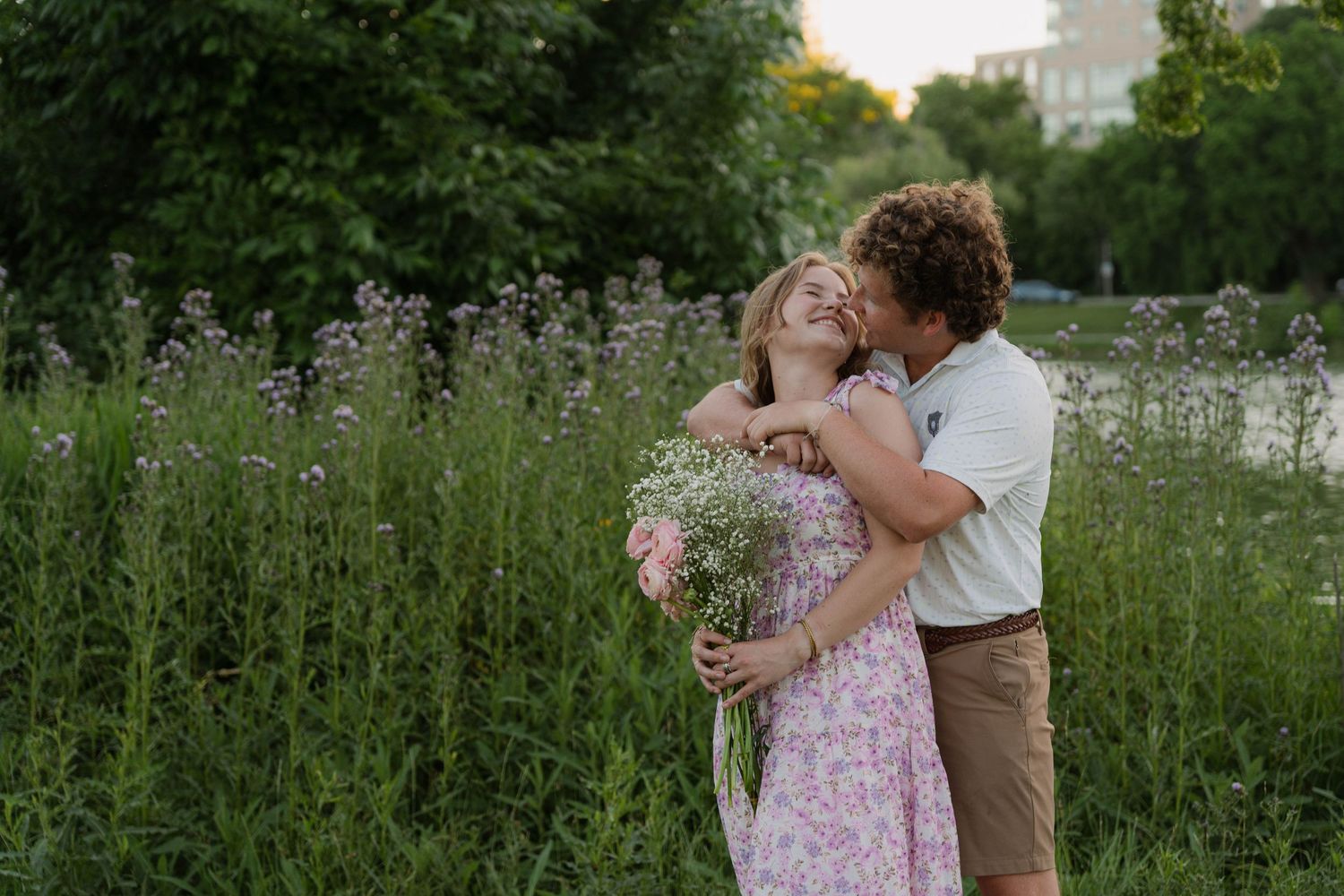 happy couple kissing by tall green grass holding green and pink boquet wearing pink dress white shirt brown pants with skyline background