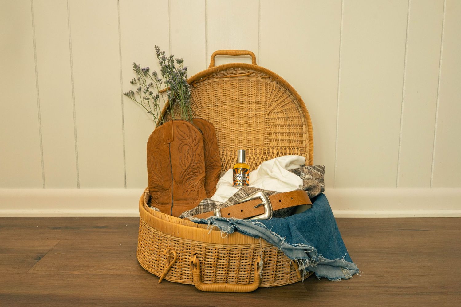 retro perfume branding photo in wicker suitcase with cowboy boots, silver buckle tan be;t, and lavender sprigs.