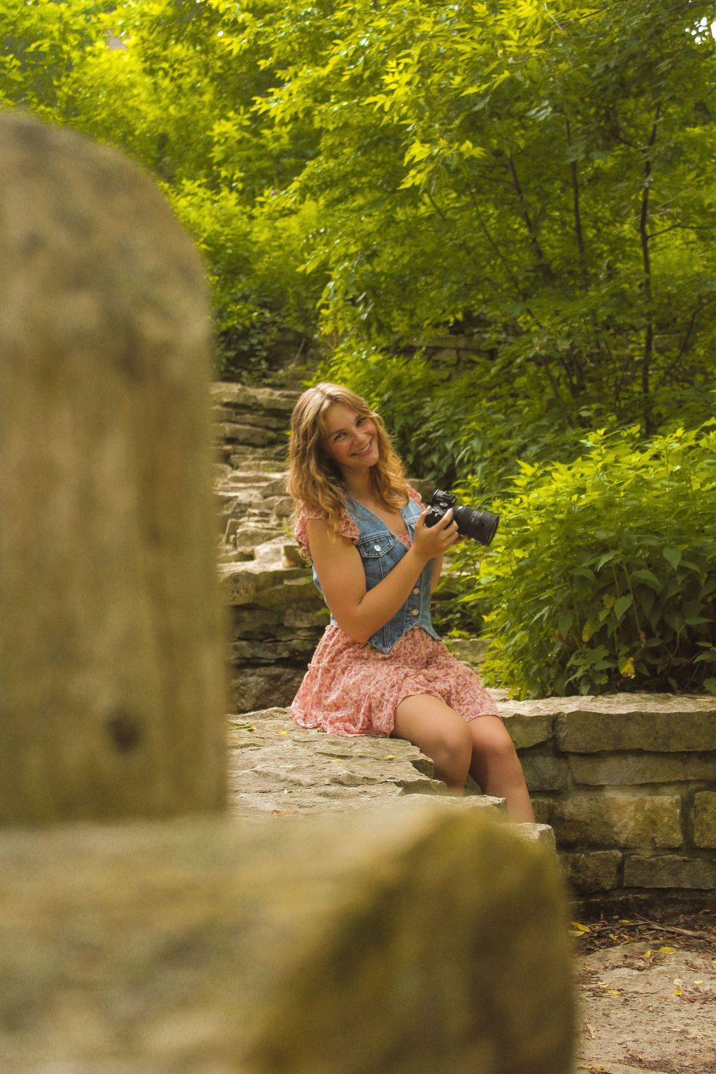 Girl photographer smiling holding camera and sitting on rocks in tall green trees wearing pink dress and blue vest in Milwaukee, Wisconsin