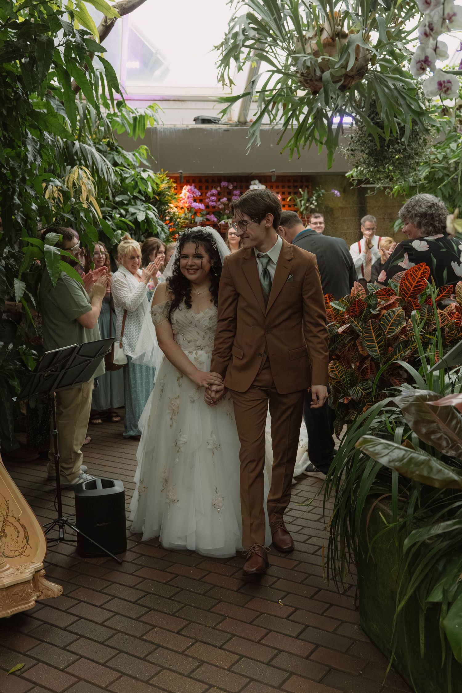 Happy bride and groom walking down brick aisle at Bolz Conservatory in Madison, Wisconsin surrounded by lush tropical greenery and family
