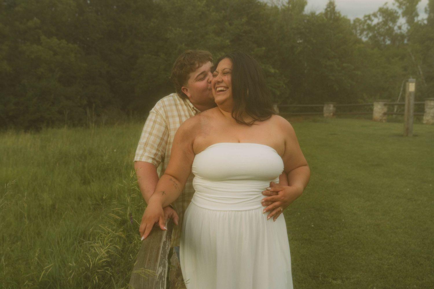 happy couple with man kissing woman on cheek at Fonferek's Glen in Denmark, Wisconsin in grassy field wearing yellow shirt and white dress