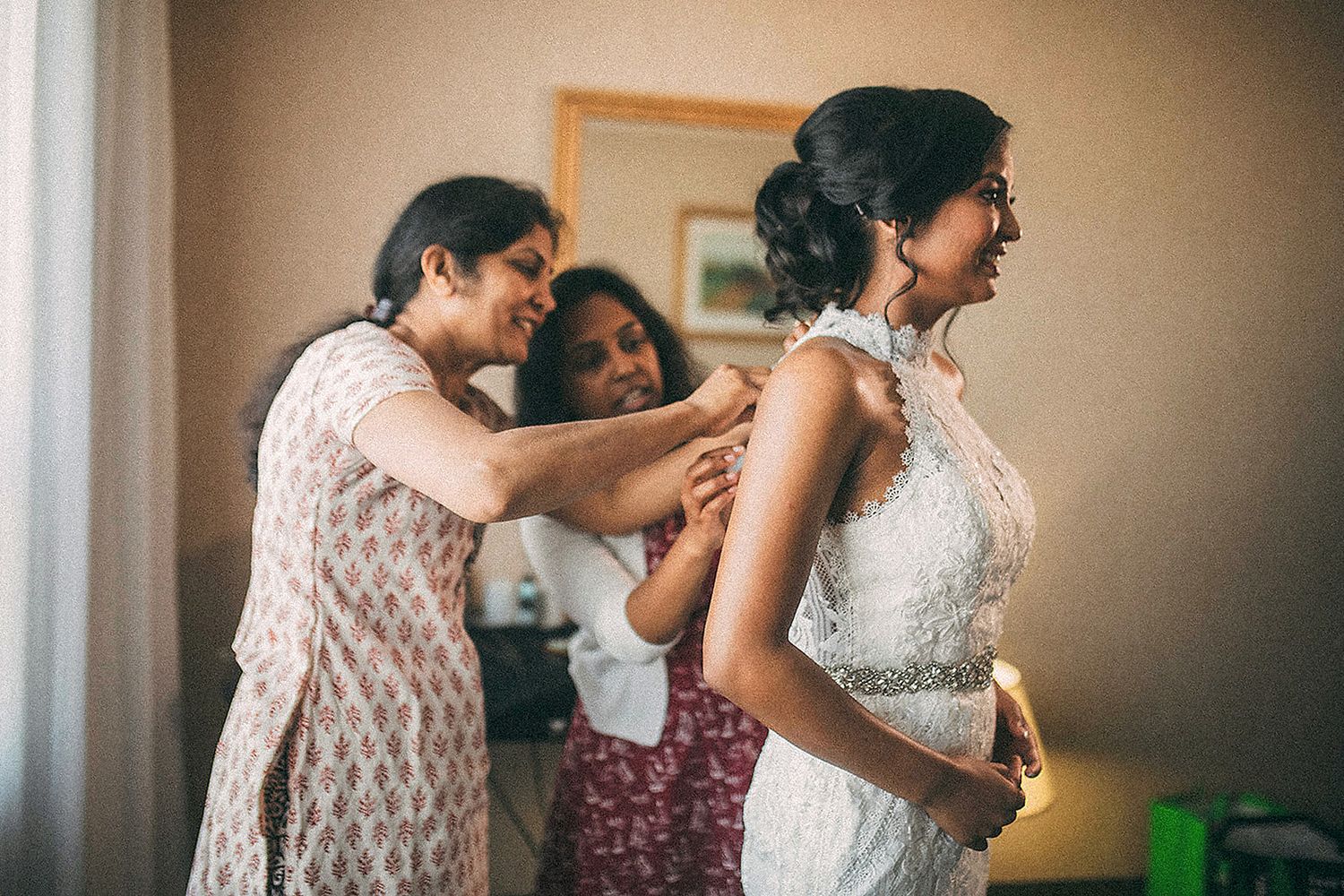Two women helping a bride in lace wedding dress with final preparations in a softly lit room.