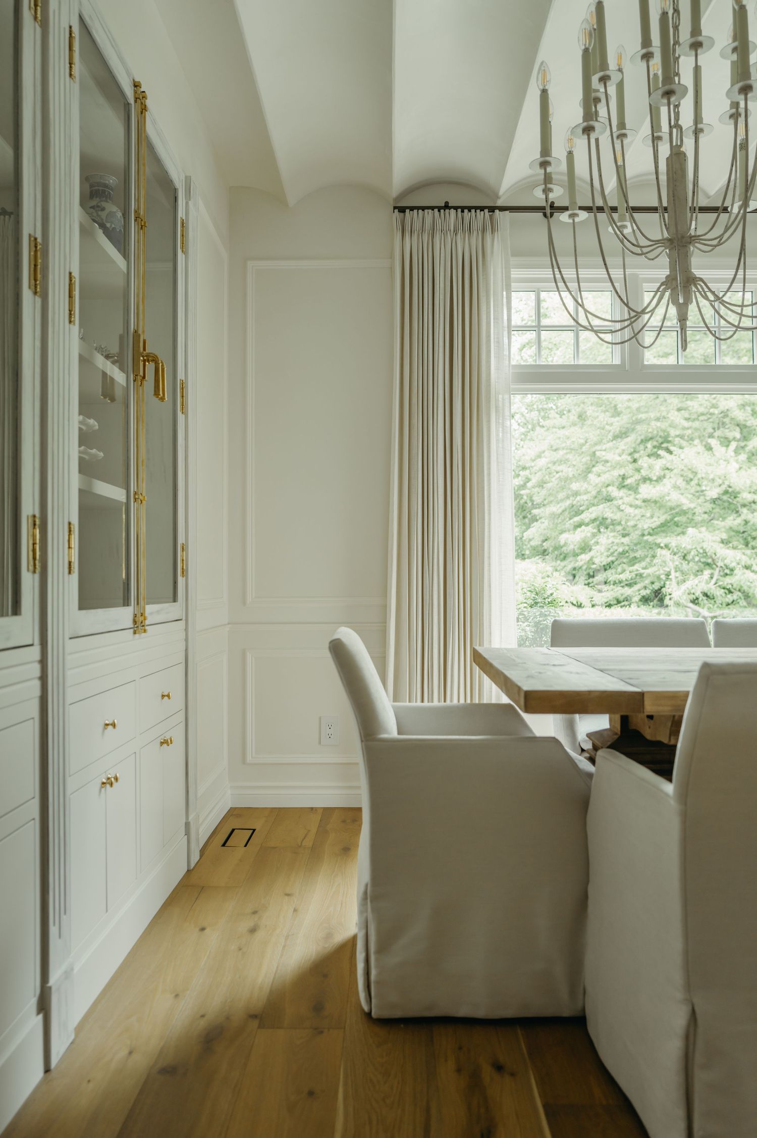 Elegant dining room with white cabinetry, gold hardware, linen curtains, slipcovered chairs, wood floors, and a beaded chandelier.