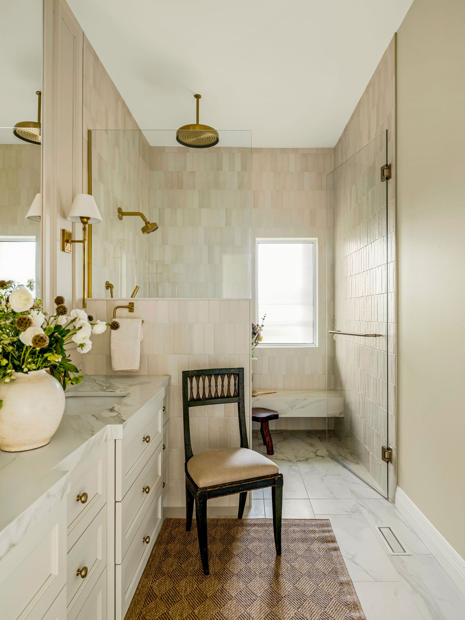 Elegant bathroom with beige tile walls, white vanity with brass fixtures, marble countertop, walk-in shower, and decorative chair.