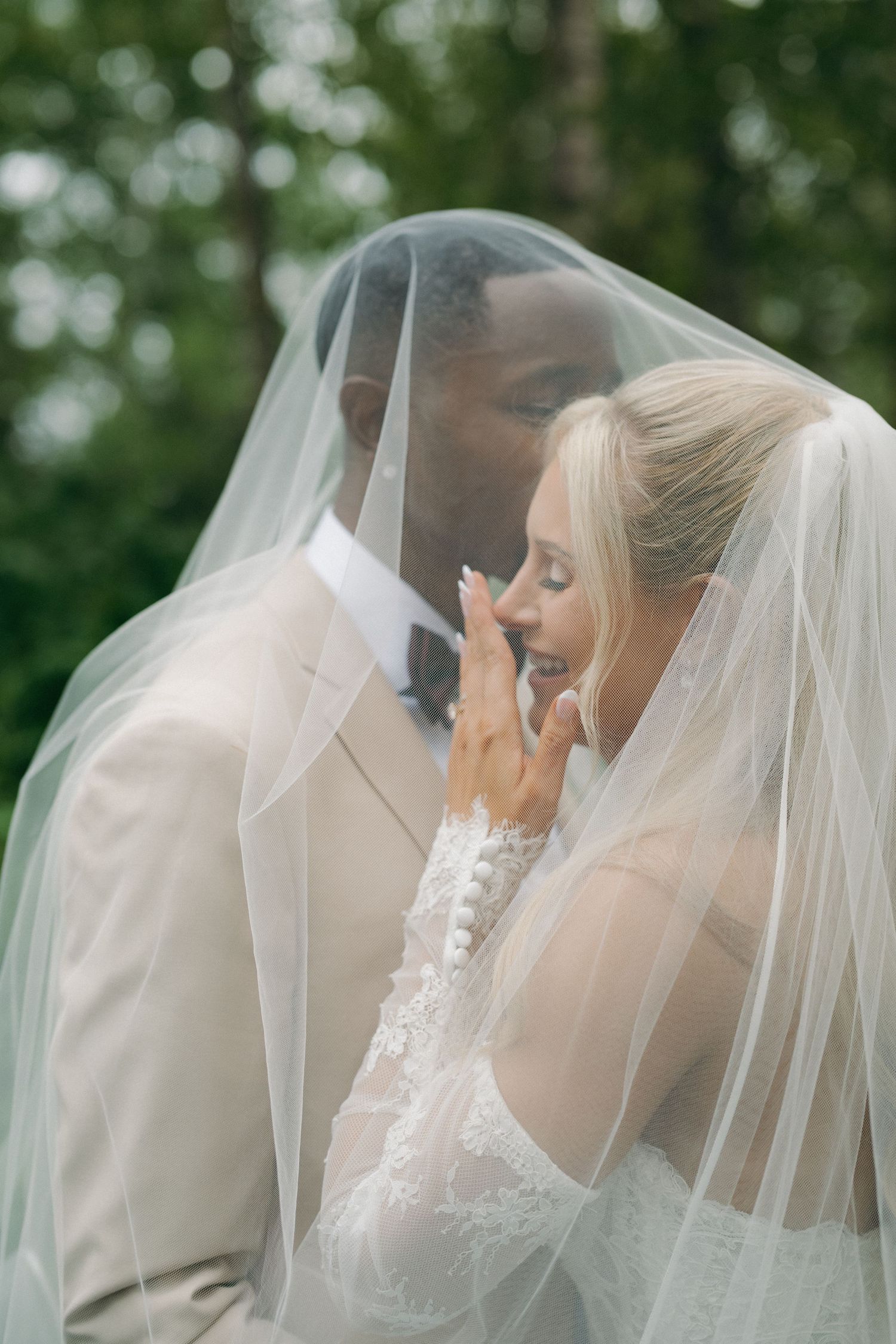 Interracial wedding couple sharing intimate moment under bride's veil outdoors.