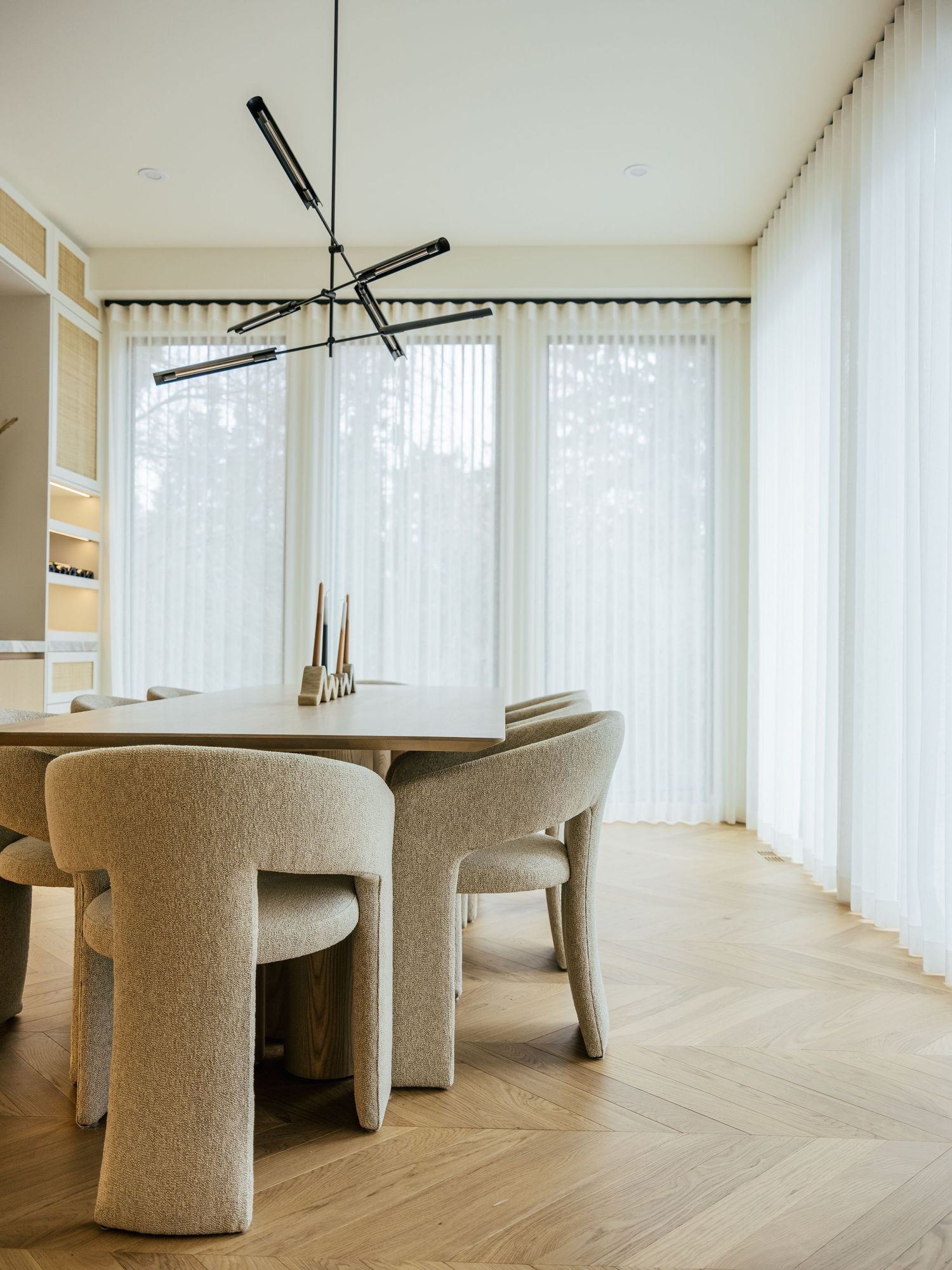 Modern dining room with beige boucle chairs, wood herringbone floor, sheer curtains, and black geometric pendant light.