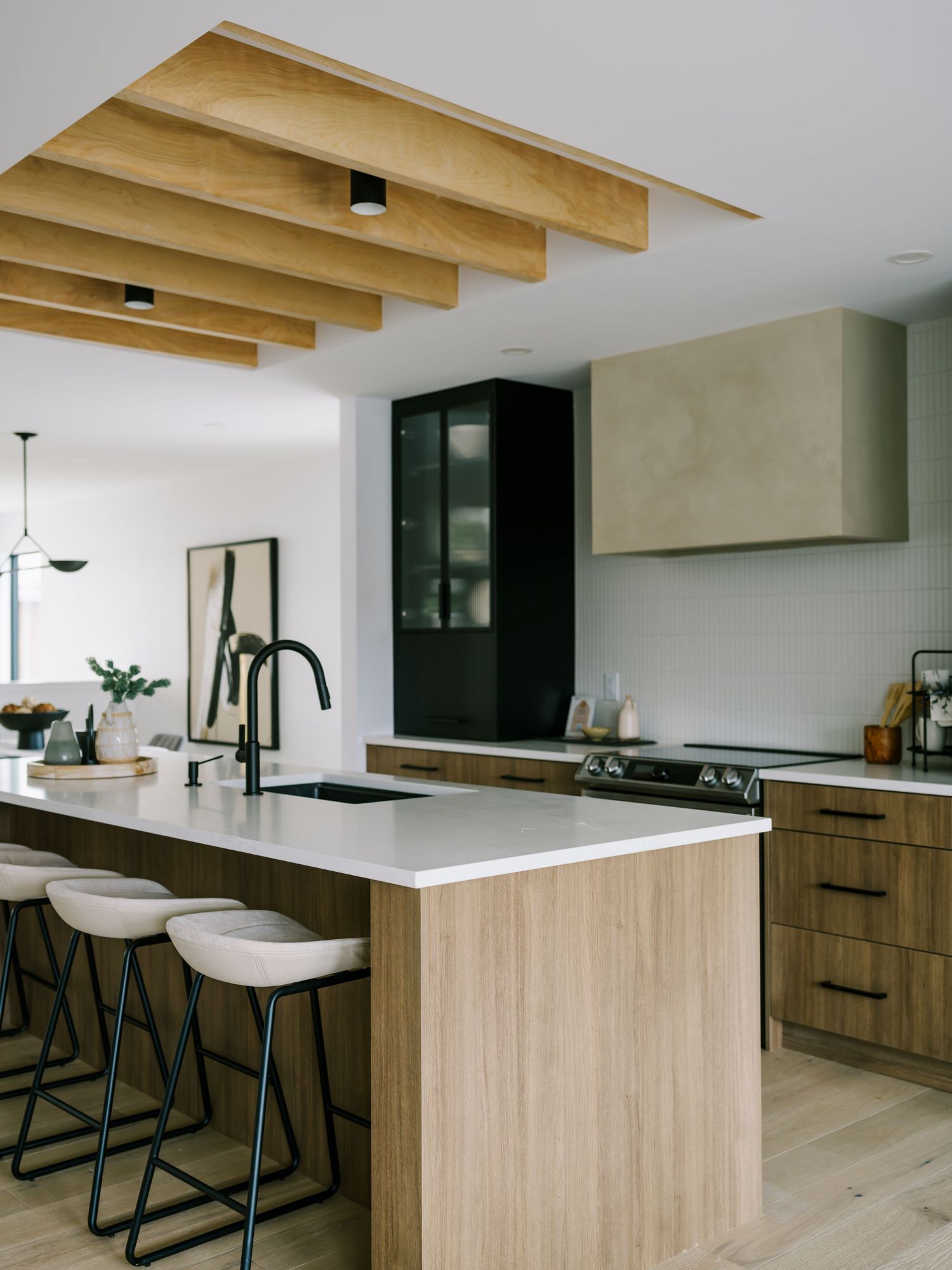 Modern kitchen with wood grain cabinets, white quartz island, bar stools, black fixtures, and exposed wood beam ceiling.