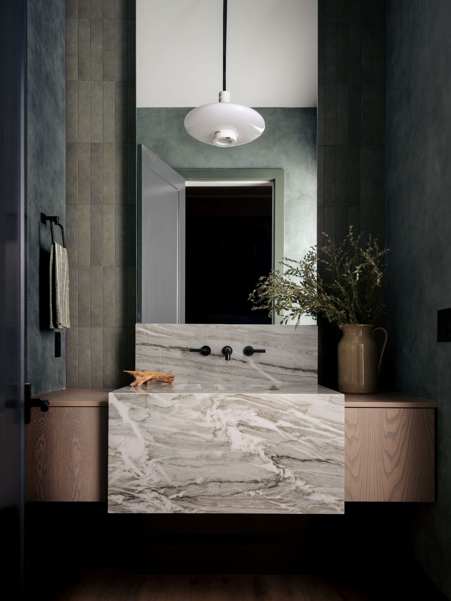 Elegant bathroom with marble vessel sink, floating wood vanity, green tiled walls, pendant light, and black wall-mount faucet.