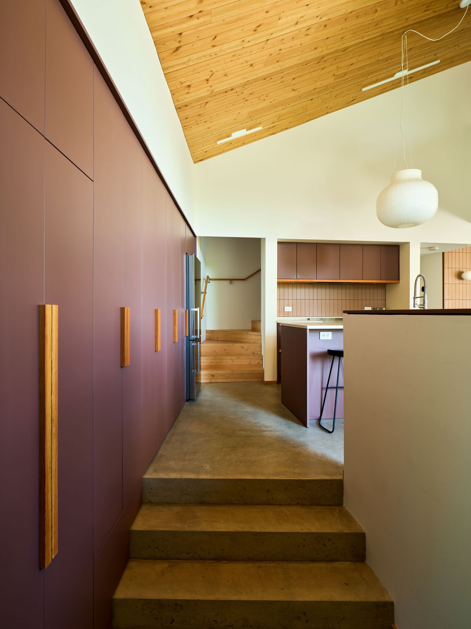 Modern hallway with mauve built-in cabinets, wood accents, concrete steps, and vaulted pine ceiling leading to open kitchen.