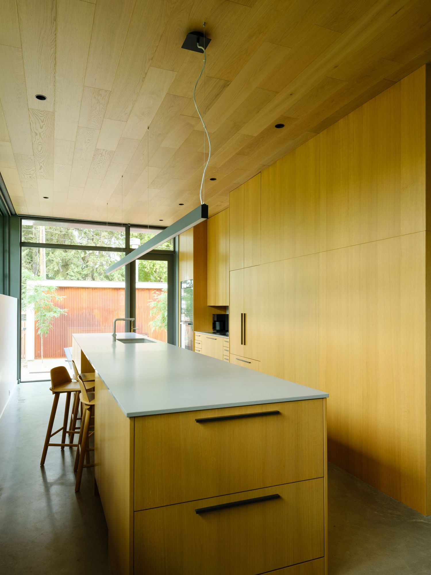 Sleek modern kitchen with wood cabinetry, gray island countertop, bar stools, and wood plank ceiling.