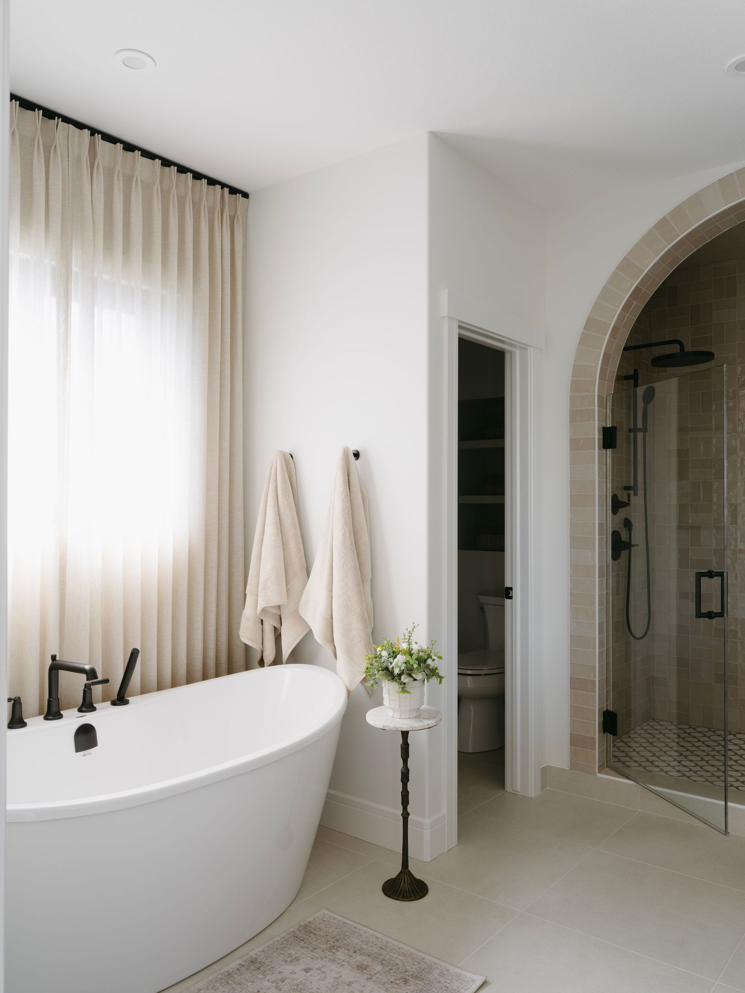 Elegant white bathroom with freestanding soaking tub, black fixtures, sheer curtains, and arched stone shower entry.