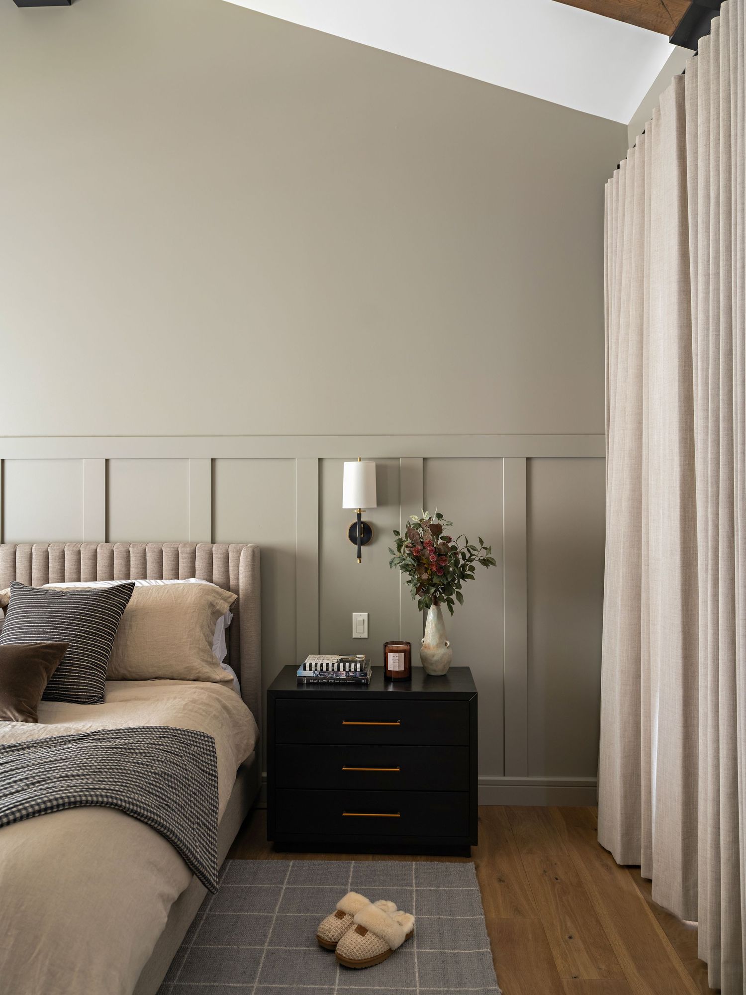 Cozy bedroom with sage green wainscoting, tufted blush headboard, black nightstand, and linen curtains.