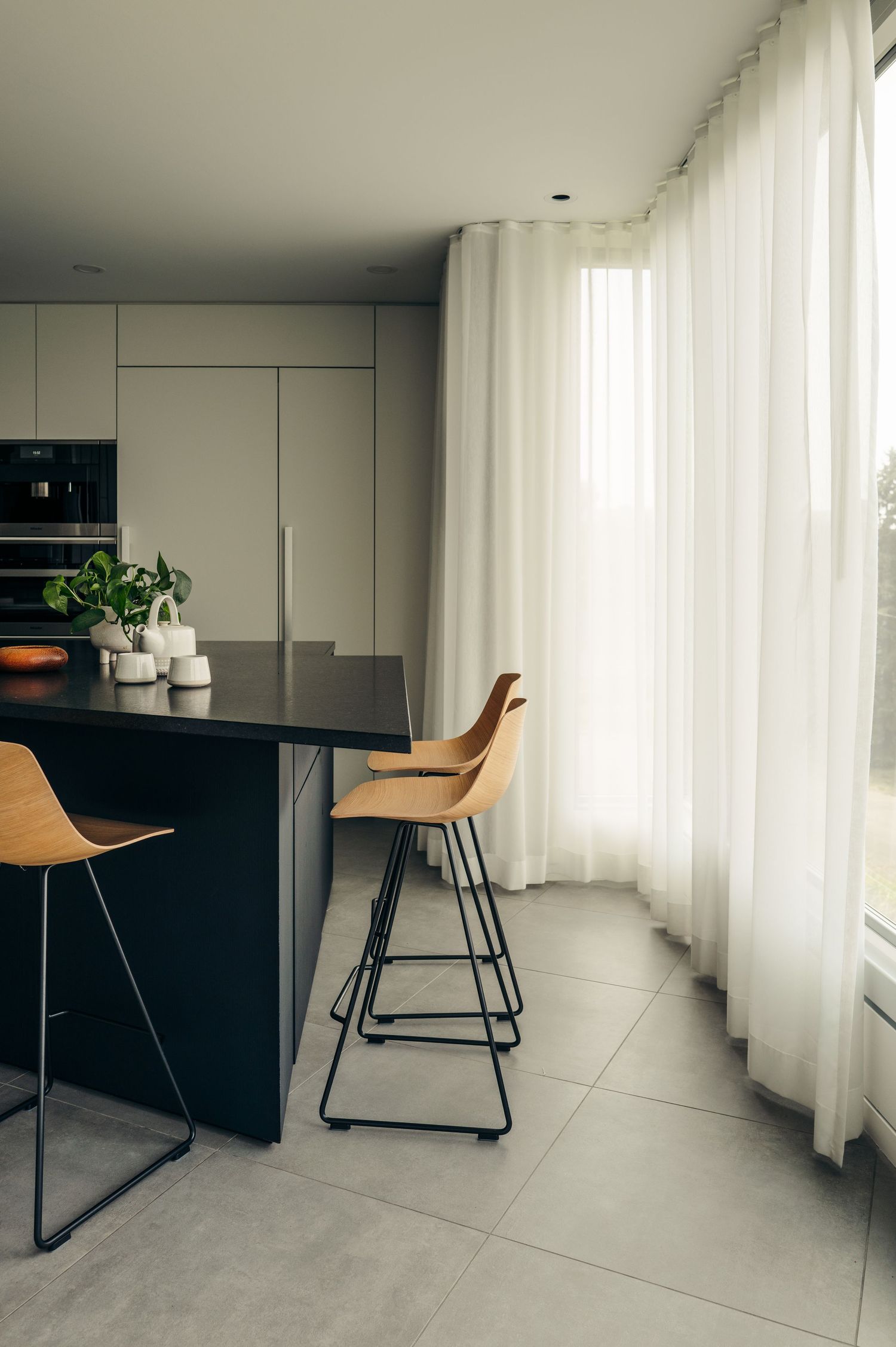 Modern kitchen with dark island, wooden bar stools, grey cabinetry, and sheer white floor-to-ceiling curtains.