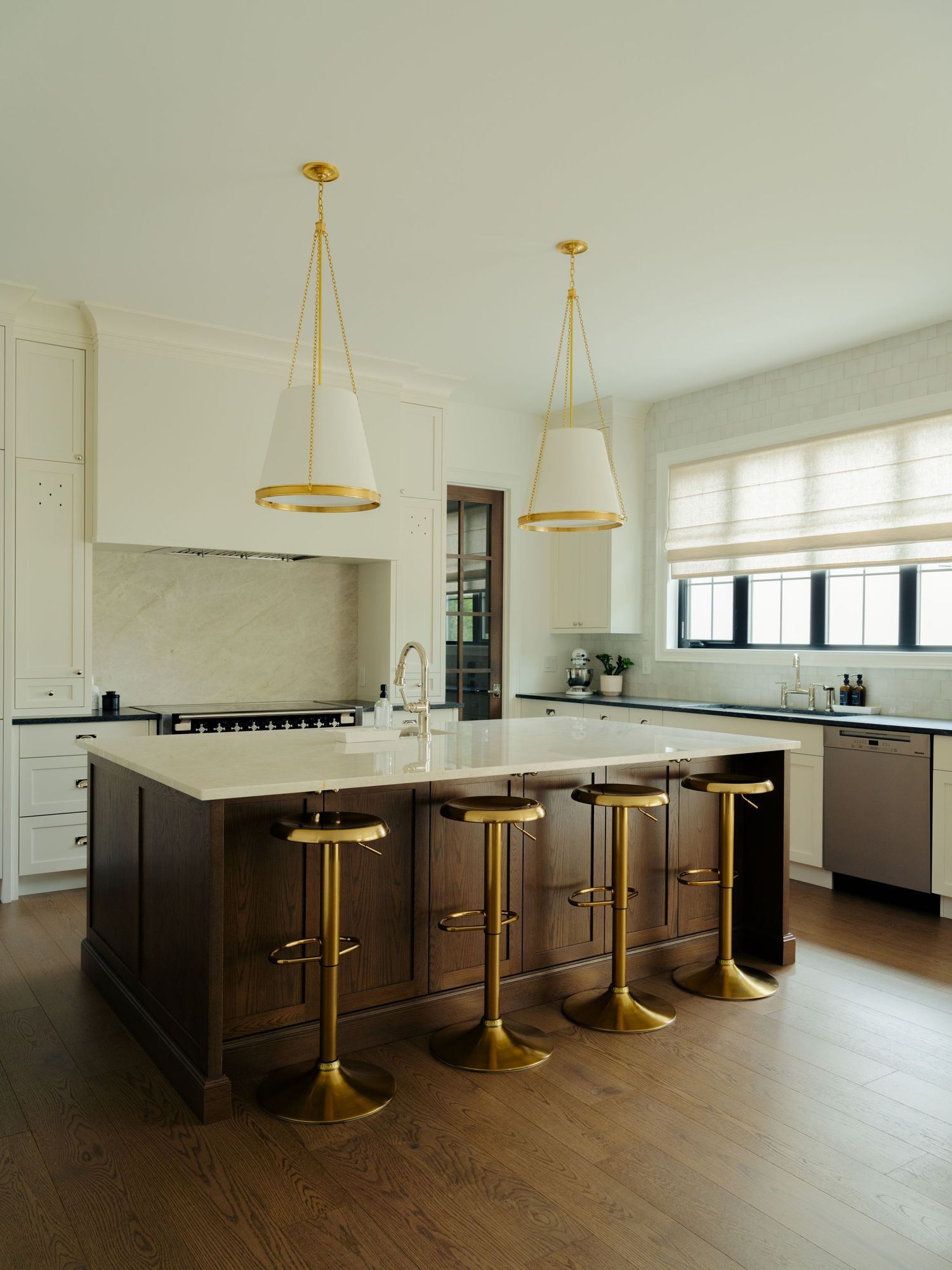 Elegant kitchen with dark wood island, gold bar stools, white pendant lights, cream cabinetry, and marble backsplash on hardwood floors.