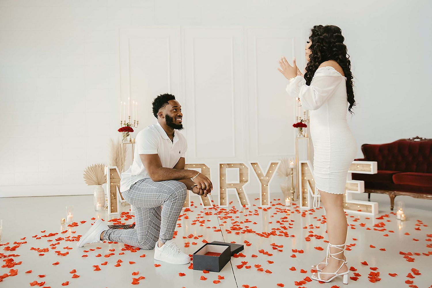 Man on one knee proposing marriage surrounded by rose petals with MARRY ME letters in background.