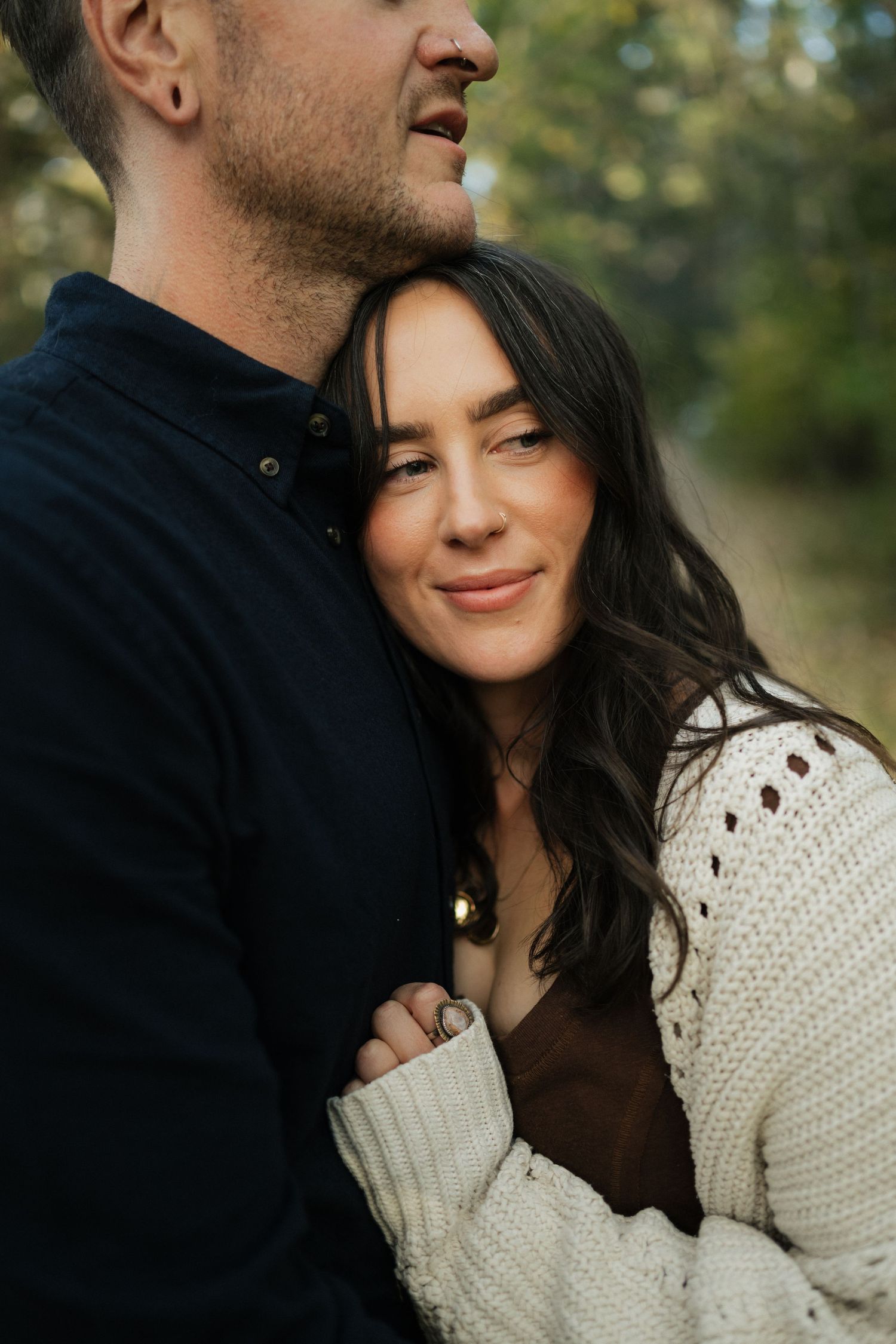 Woman in a cream knit sweater smiles while leaning against a man in a navy shirt in an autumn outdoor setting.