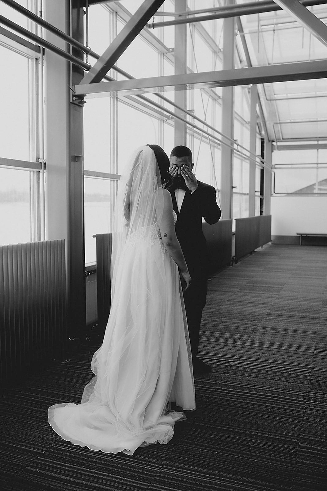Black and white photo of groom wiping tears during emotional first look with bride in veil.