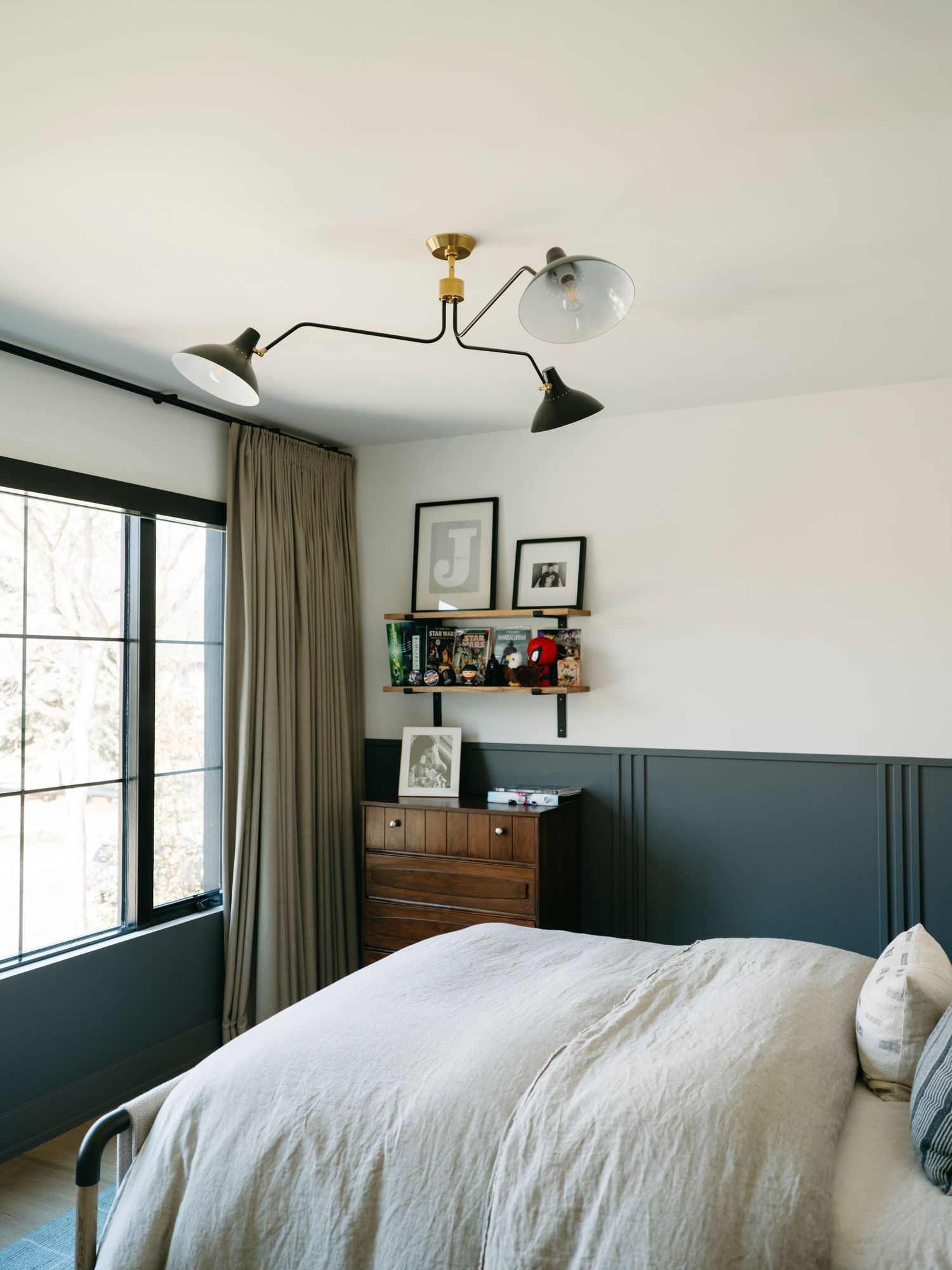 Bedroom with dark wainscoting, mid-century ceiling light, wood dresser, and wall shelf with decorative items.
