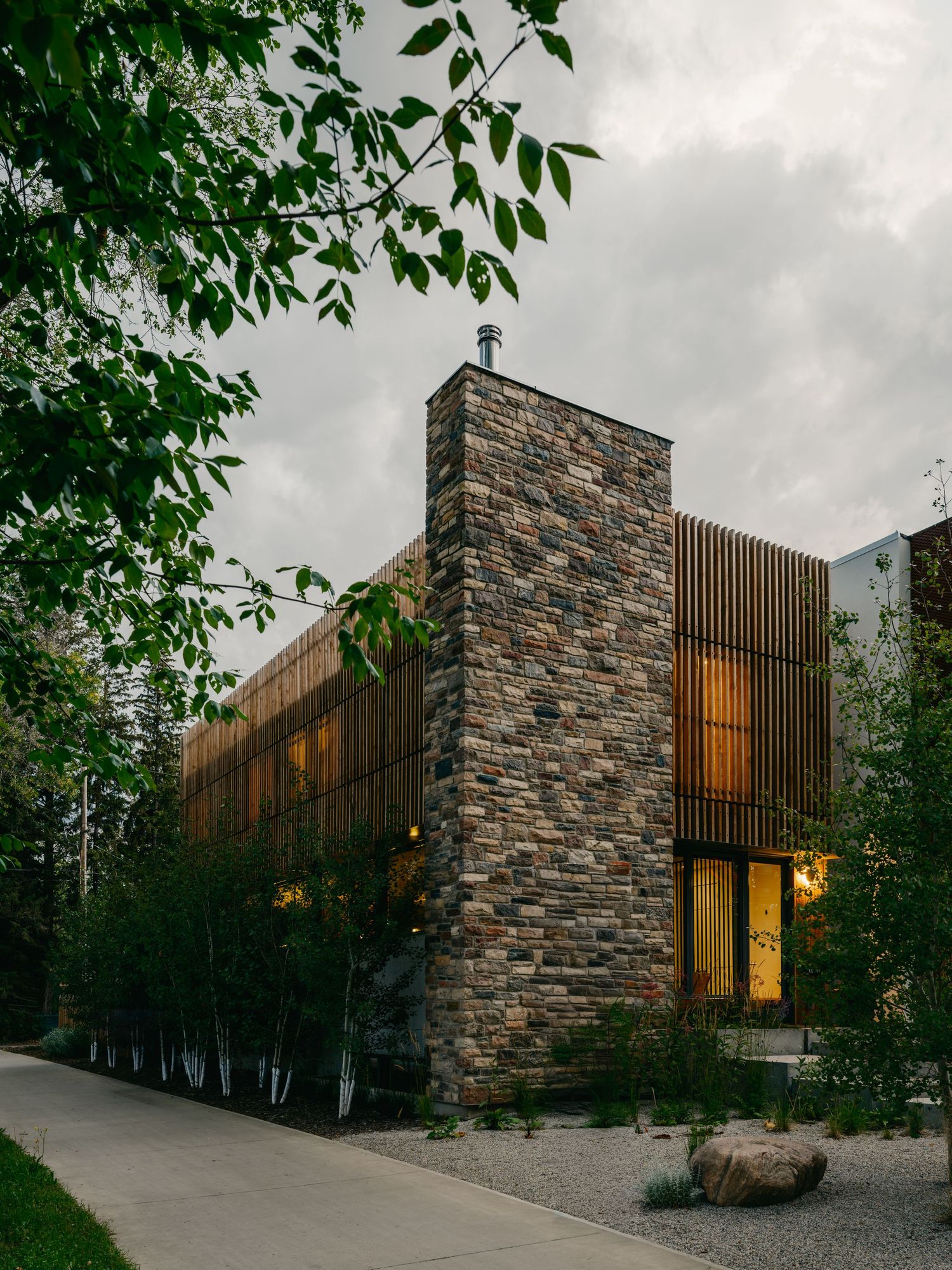 Contemporary stone and wood exterior home facade surrounded by lush green trees at dusk.