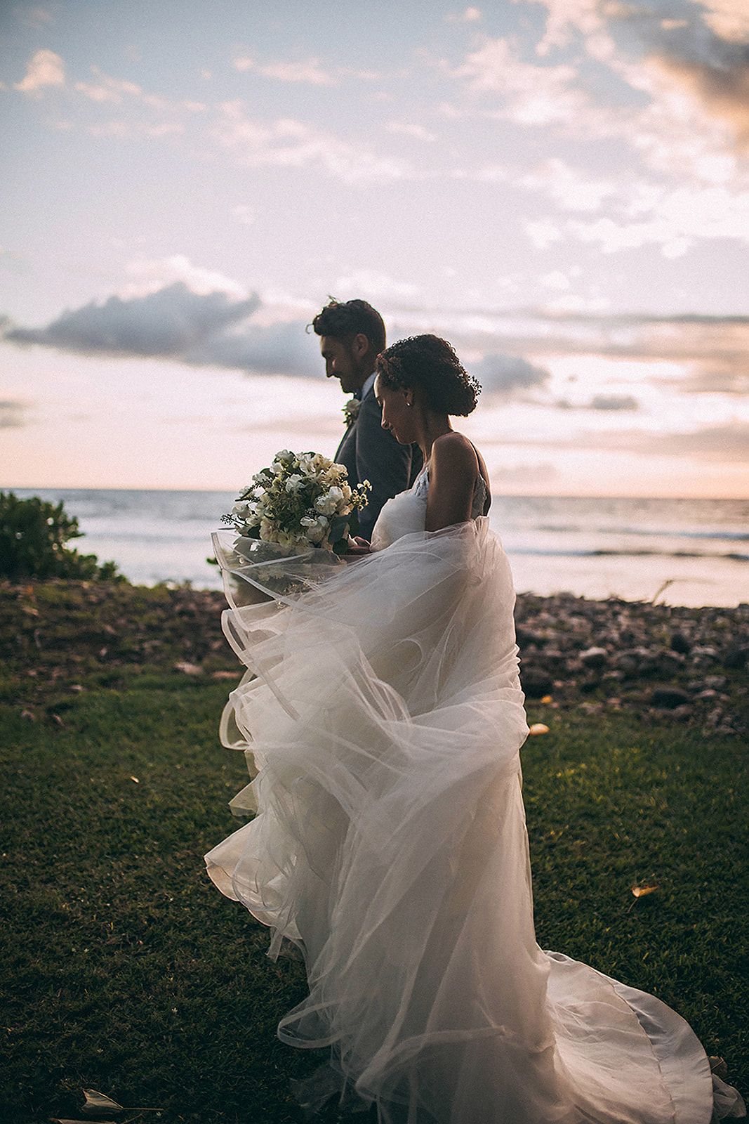 Bride and groom standing by the ocean at sunset, her layered tulle gown flowing in the breeze.