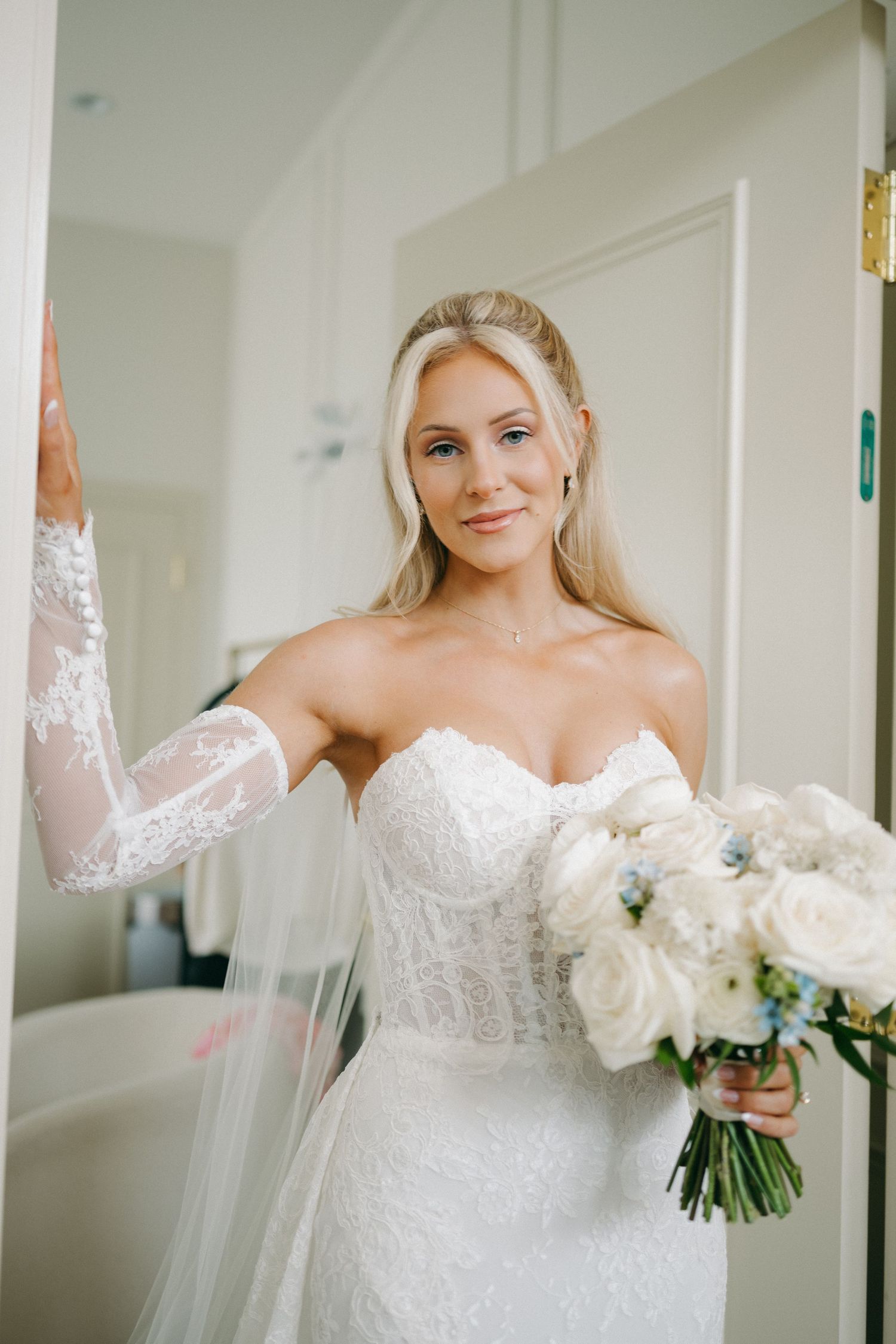 Blonde bride in strapless lace gown holding white and blue bouquet posing in doorway.