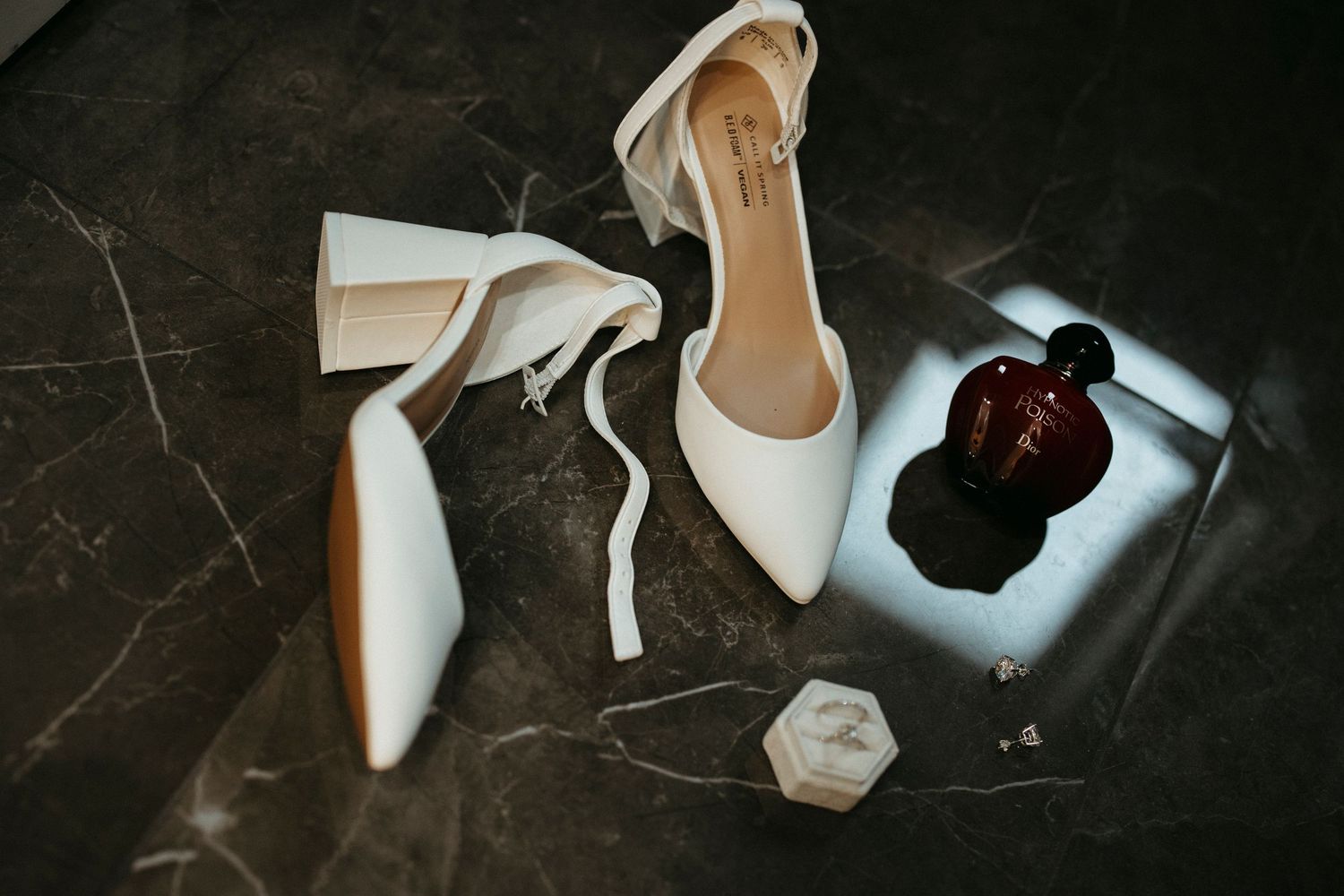 White bridal heels with ankle ties displayed on dark marble surface beside perfume and ring box.