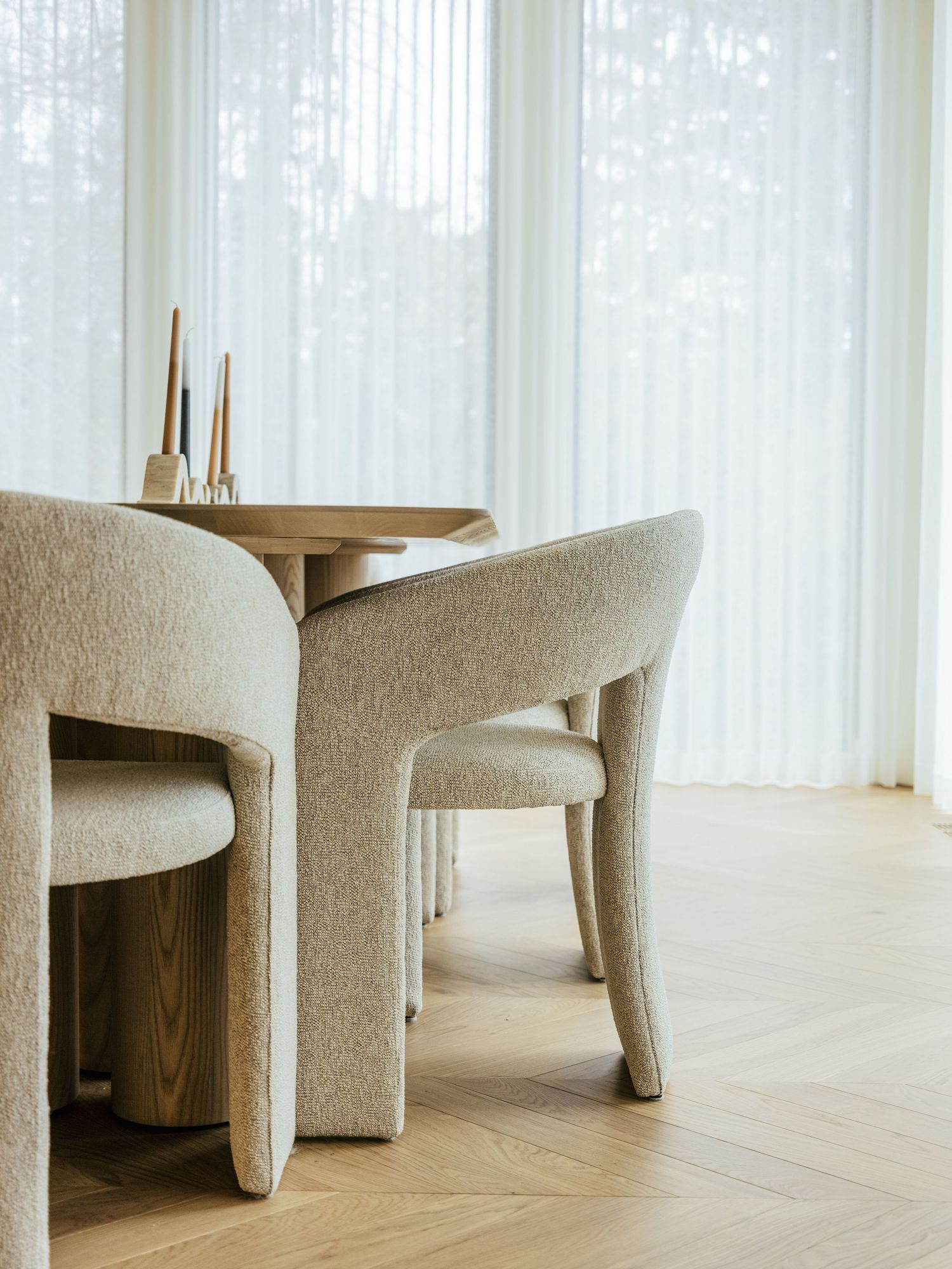 Minimalist dining room with bouclé chairs and round wooden table on herringbone oak floors with sheer white curtains.