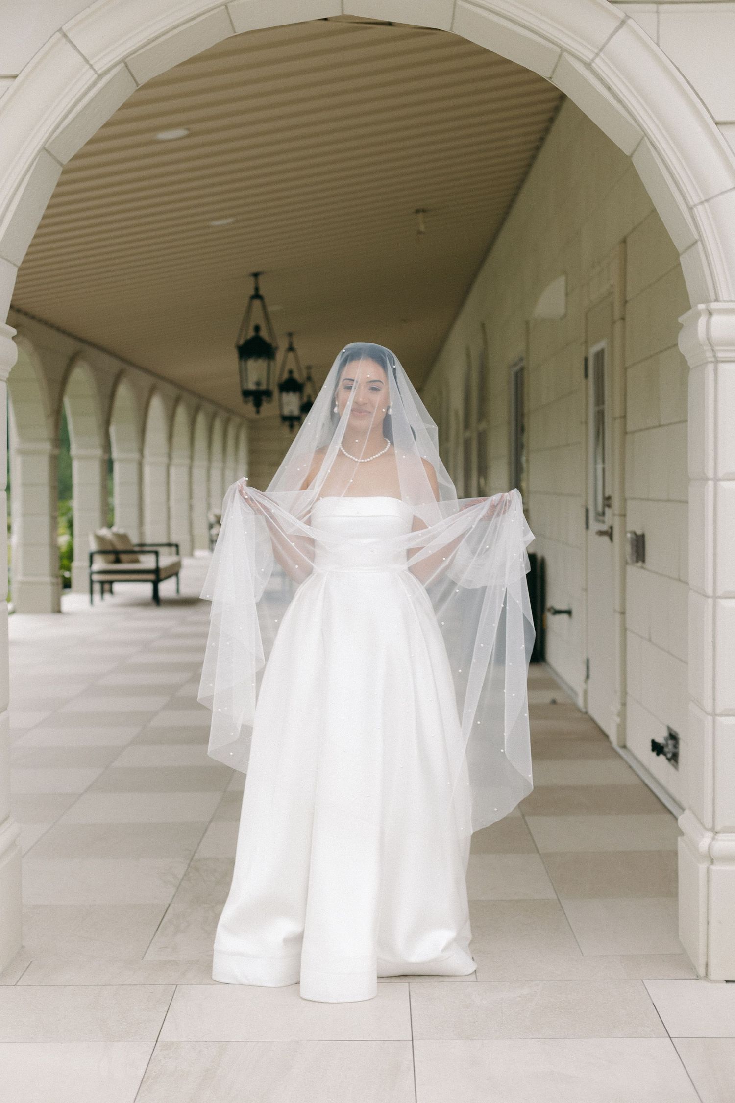Bride in strapless white wedding dress holds flowing veil wide while walking through arched stone corridor.