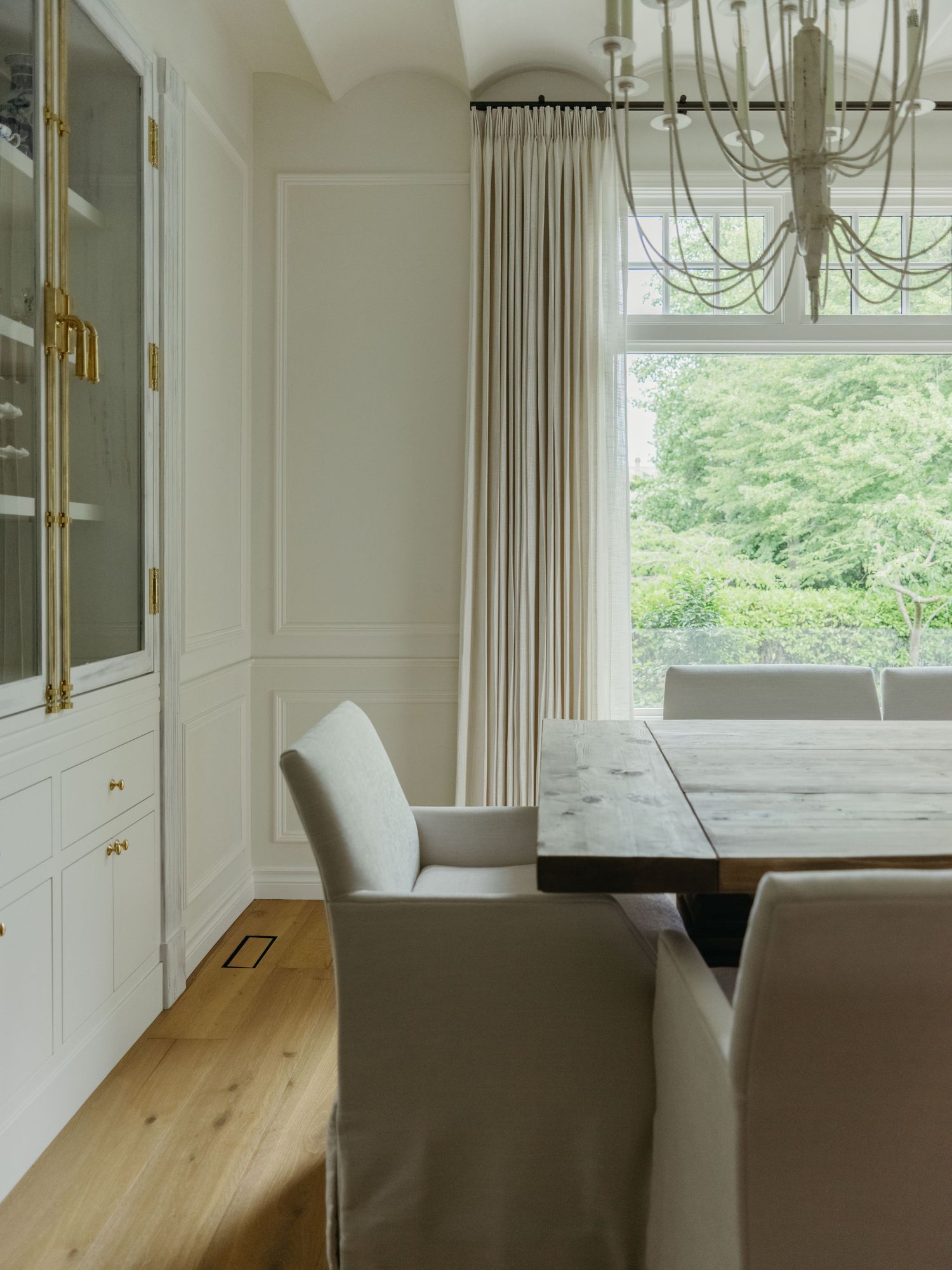 Elegant dining room with white wainscoting, brass cabinet, marble table, slipcovered chairs, linen drapes, and gold chandelier.